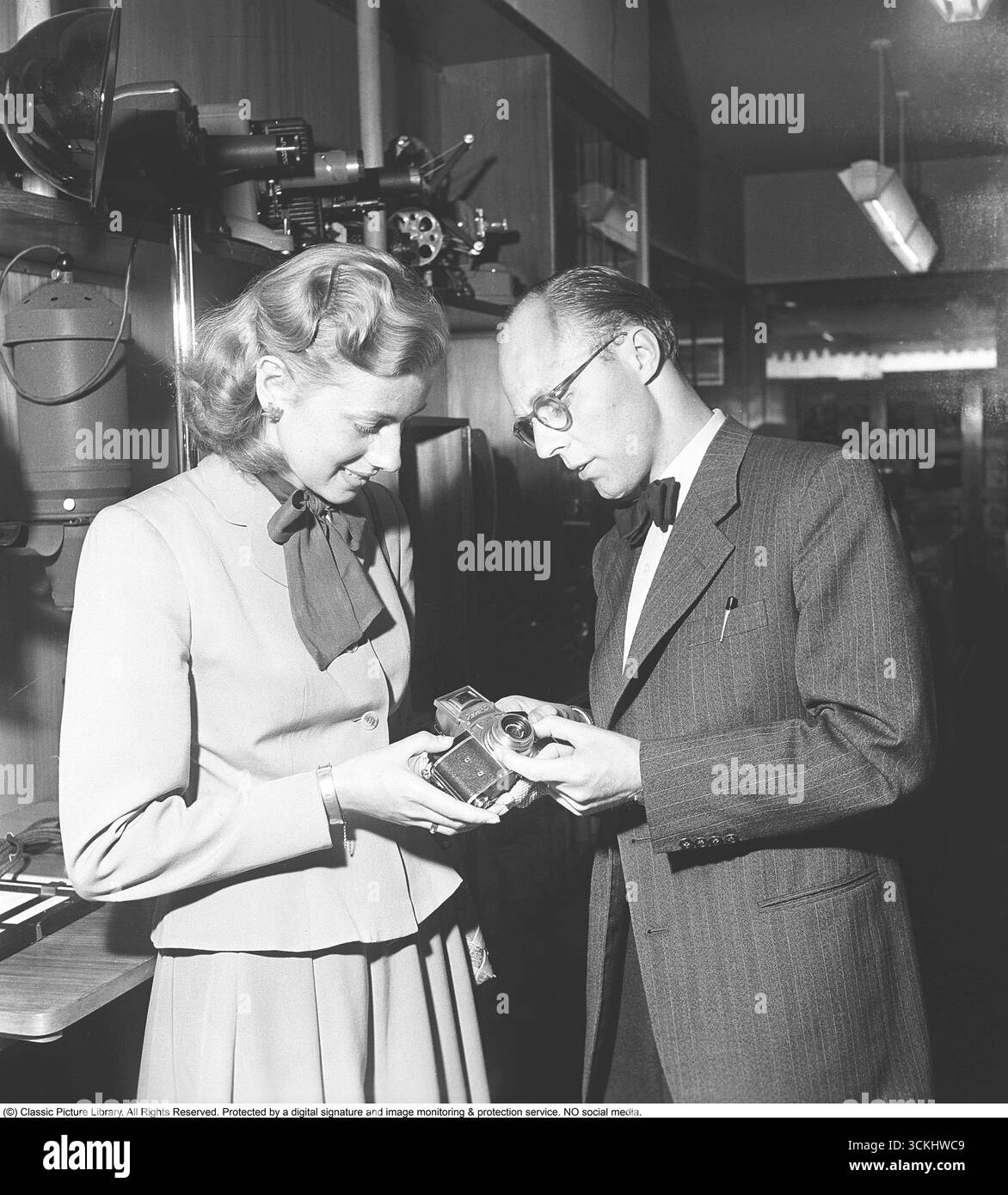 In un negozio di macchine fotografiche e attrezzature fotografiche nel 1949, a una giovane donna viene mostrata la fotocamera Exakta II. La Exakta II, introdotta nel 1949 da Ihagee Kamerawerk a Dresda, era una fotocamera reflex a obiettivo singolo compatta da 35 mm, basata sul design pre-bellico di Kine Exakta. Prodotto in quantità limitate sotto il controllo sovietico dopo la seconda guerra mondiale, era dotato di un otturatore a sinistra, mirino a livello della vita e compatibilità con gli obiettivi Zeiss Jena e Meyer premium. Quell'anno, un accessorio pentapristico ha consentito una visione a livello degli occhi, aumentando il suo fascino. Contrassegnato come "prodotto in Germania", è diventato un'importazione ricercata in Foto Stock