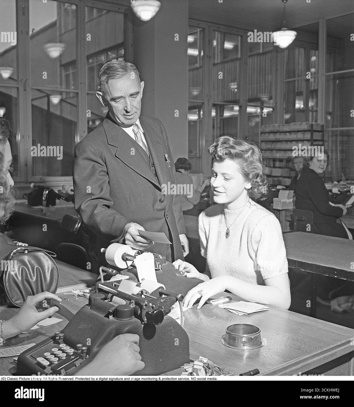 Un ufficio bancario negli anni '1940 Le lavoratrici lavorano presso la cassa di risparmio postale, registrando pagamenti, prelievi e depositi. Un uomo sta accanto a un impiegato di una calcolatrice meccanica, leggendo entrambi da un piccolo foglio di carta. Questa scena riflette il crescente ruolo delle donne nel settore bancario e l'uso della tecnologia d'ufficio precoce nell'Europa del dopoguerra. Gli anni '1940 hanno visto un aumento delle donne che entrano nella forza lavoro, tra cui le banche, mentre gli sforzi di recupero post-guerra hanno ampliato i ruoli clericali. Le banche di risparmio postale, diffuse in Europa e oltre, fornivano servizi finanziari accessibili. (Kristoffersson, rif. AU7 Foto Stock