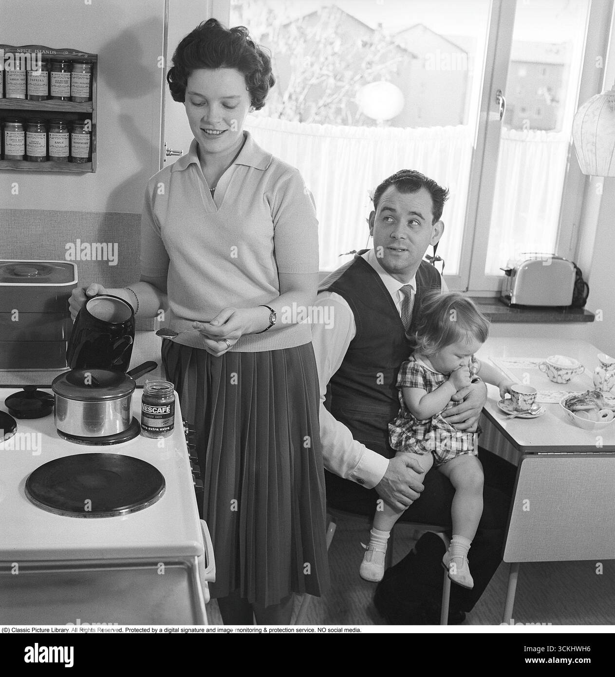 Prepara il caffè nel 1961. Una coppia nella loro cucina, dove la moglie usa il nuovo marchio Nescafé per preparare un delizioso caffè per suo marito. Le tazze da caffè sono disposte sul tavolo della cucina, con il motivo Virrvarr disegnato da Sigvard Bernadotte. Questa scena riflette l'ascesa del caffè istantaneo e il design scandinavo della metà del secolo nelle famiglie del dopoguerra. Gli anni '1960 videro l'ascesa globale del caffè istantaneo, con Nescafé (introdotto da Nestlé nel 1938) che guadagnò popolarità per la sua convenienza, specialmente nell'Europa del dopoguerra, dove furono abbracciati i moderni elettrodomestici e prodotti da cucina. (Kristoffersson, Foto Stock