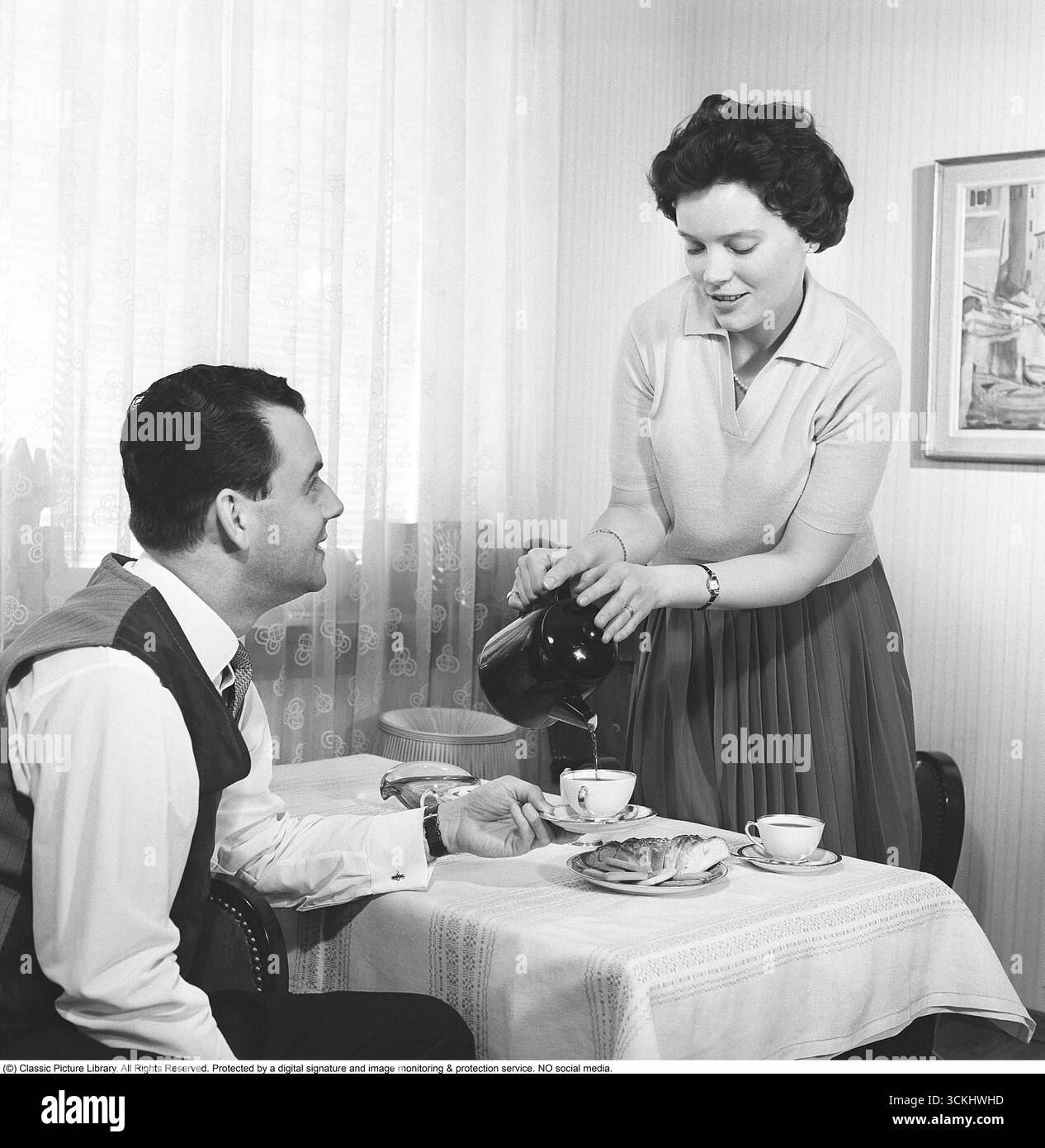 Versa il caffè nel 1961. Una coppia al loro tavolo da cucina dove la moglie serve il nuovo marchio Nescafé per preparare un delizioso caffè istantaneo per suo marito. Le tazze da caffè sono disposte sul tavolo della cucina con panini e biscotti. Questa scena riflette l'ascesa del caffè istantaneo e il design scandinavo della metà del secolo nelle famiglie del dopoguerra. Gli anni '1960 videro l'ascesa globale del caffè istantaneo, con Nescafé (introdotto da Nestlé nel 1938) che guadagnò popolarità per la sua convenienza, specialmente nell'Europa del dopoguerra, dove furono abbracciati i moderni elettrodomestici e prodotti da cucina. (Kristoffersson, rif. CT14-7) Foto Stock