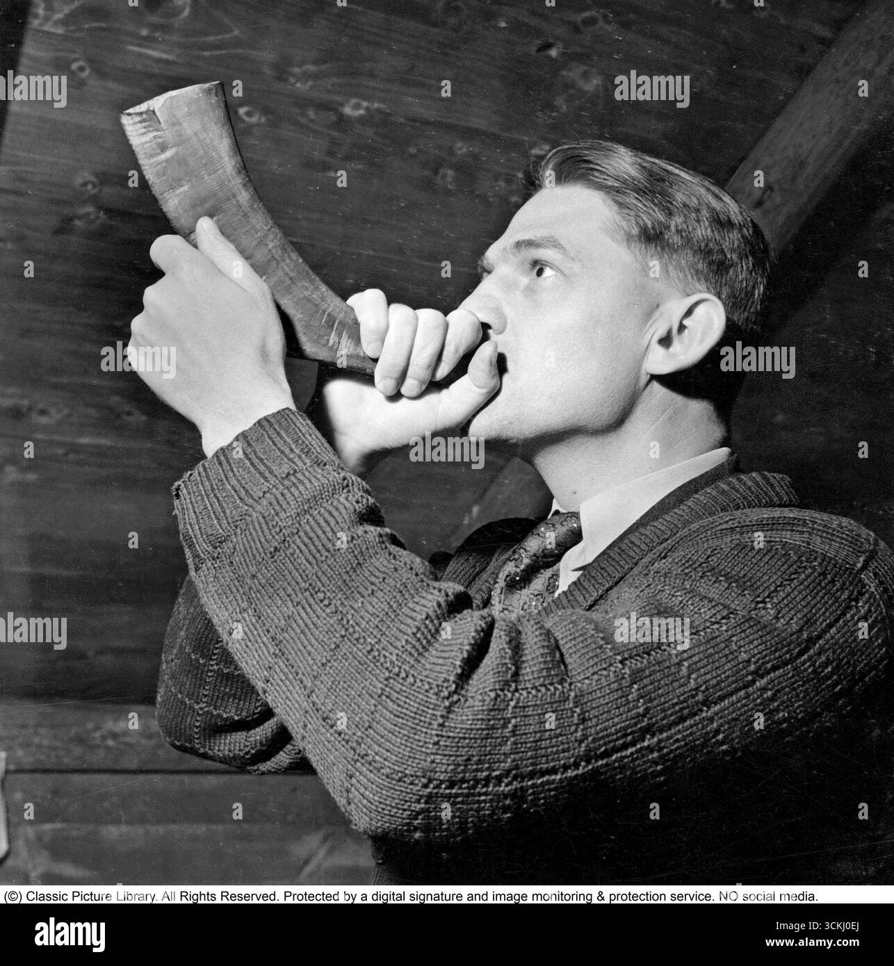 Si vede un uomo soffiare in un corno realizzato con il corno di un animale, producendo un suono profondo e risonante. Il corno, probabilmente costituito dal corno ricurvo, scavato di un ariete o di una bestia simile, è tenuto saldamente alle labbra mentre espira con forza controllata. La sua postura è stabile, le guance si gonfiano leggermente e i suoi occhi sono concentrati, mentre il tono primitivo dell'antico strumento riecheggia attraverso l'aria, evocando un senso di tradizione o cerimonia. È autore di Sven Edvin Salje (1914-1998) Foto Stock