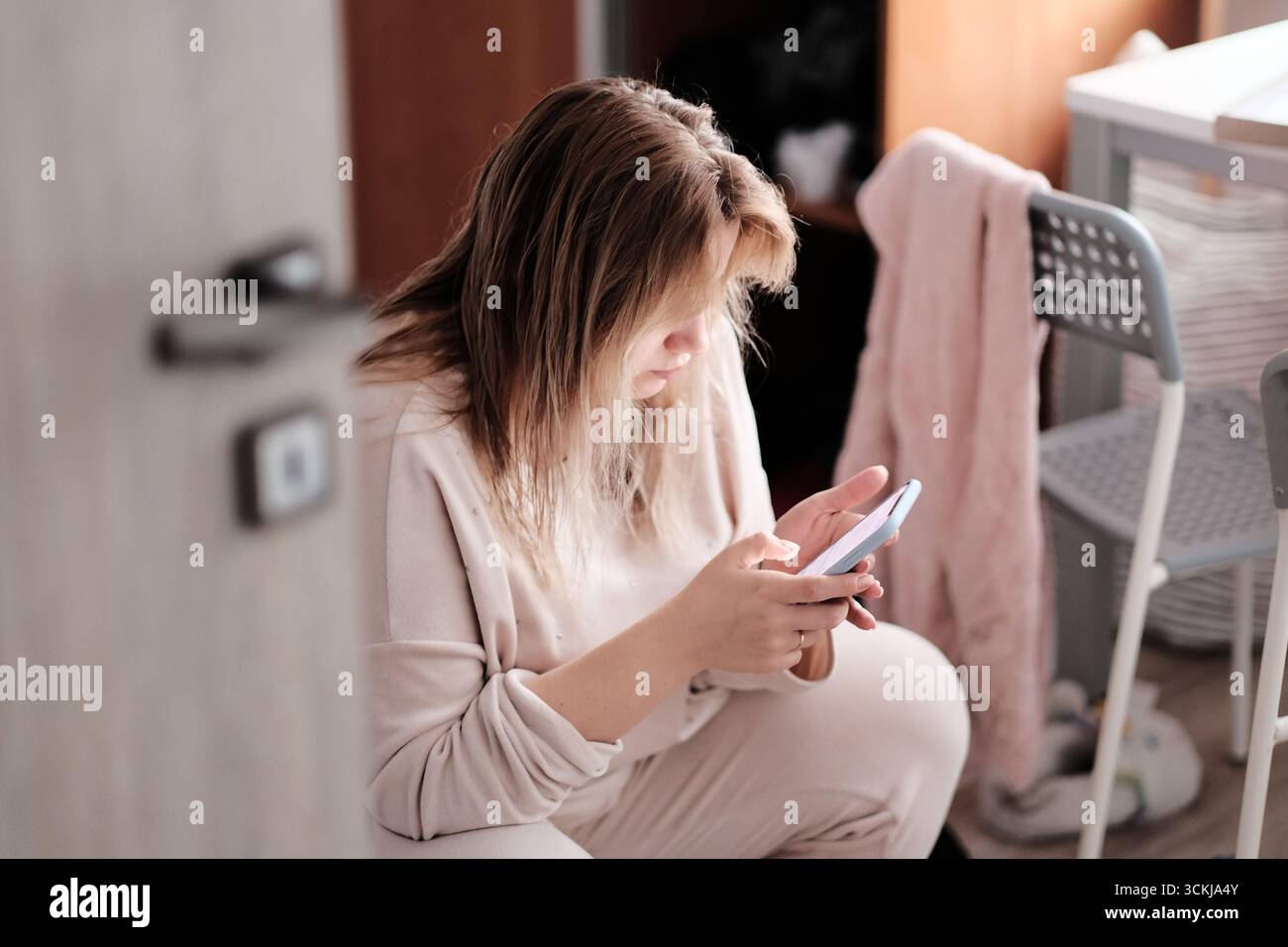 Giovane donna seduta al chiuso con un abbigliamento casual, concentrata sul suo smartphone, che trascorre il tempo online e si rilassa a casa. Foto Stock