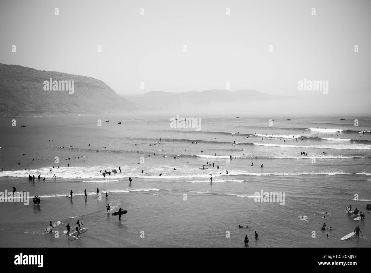 I surfisti e i nuotatori si godono le onde su una spiaggia vivace, con montagne in lontananza nebbiosa e barche che fluttuano tranquillamente all'orizzonte. Foto Stock