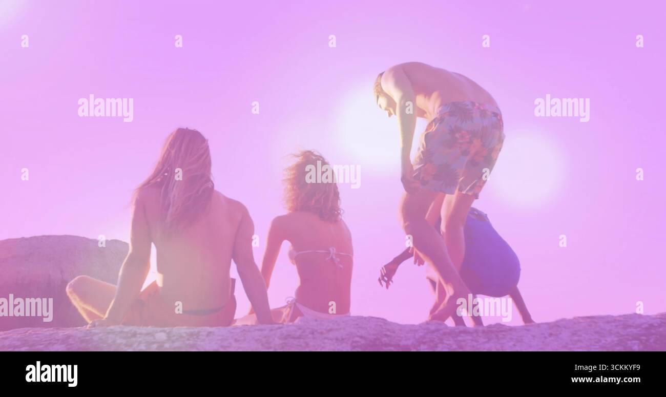 Seduti e arrampicati su quattro amici in costume da bagno su un affioramento roccioso al tramonto sulla spiaggia, con l'acqua dell'oceano Foto Stock