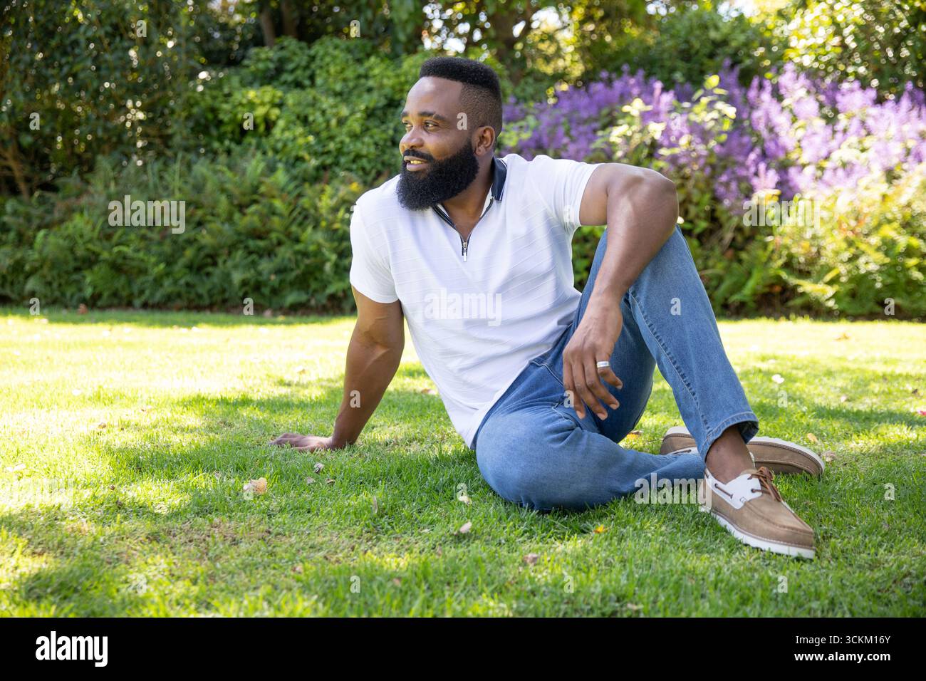 Uomo afroamericano seduto sull'erba nel parco, indossa polo bianca e jeans con anello d'argento Foto Stock