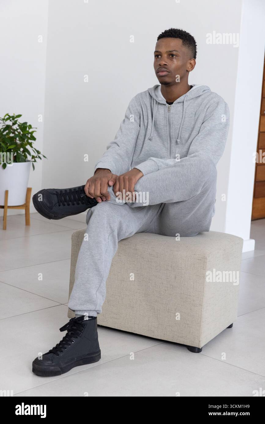 Uomo afro-americano seduto su un ottomano a casa con felpa con cappuccio grigia e sneakers nere Foto Stock