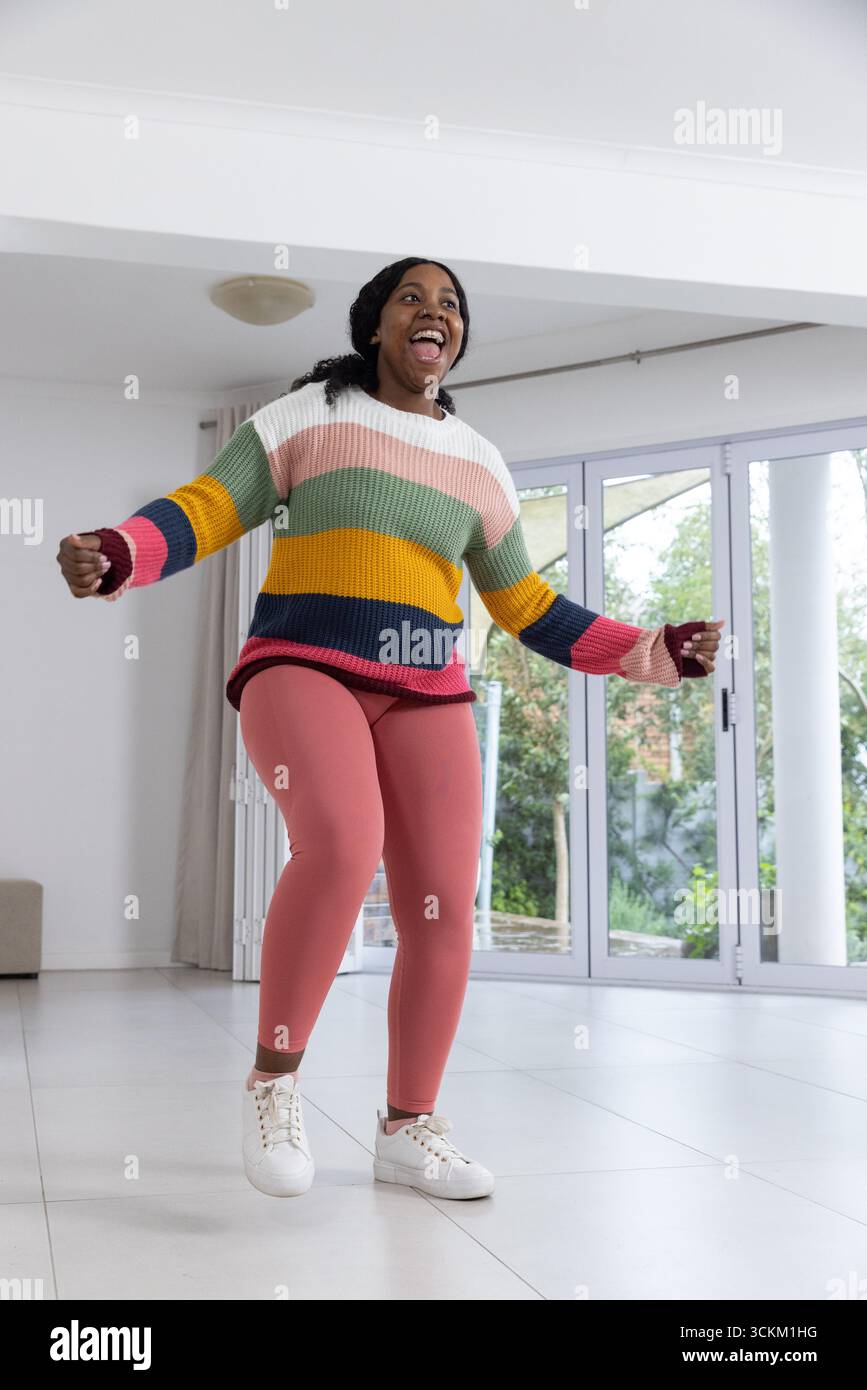 Donna afroamericana che salta nel soggiorno accanto alle porte del patio con maglione e scarpe da ginnastica Foto Stock