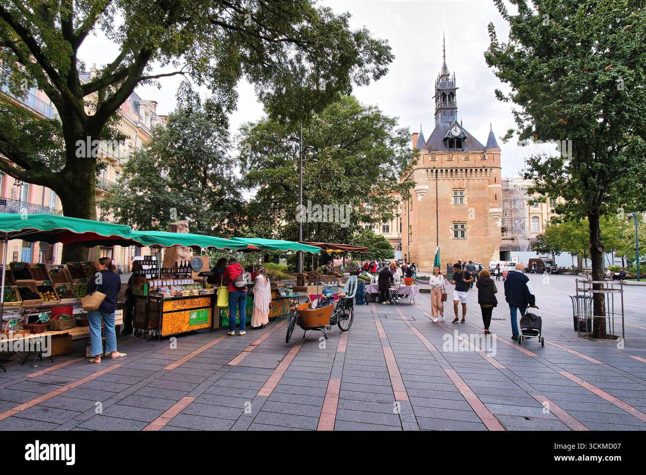 Toulouse Haute-Garonne Francia 10.09.25 piazza del mercato. Bancarelle che vendono verdure. uomini e donne che fanno shopping. Torre dell'orologio in mattoni con guglia. edificio storico Foto Stock