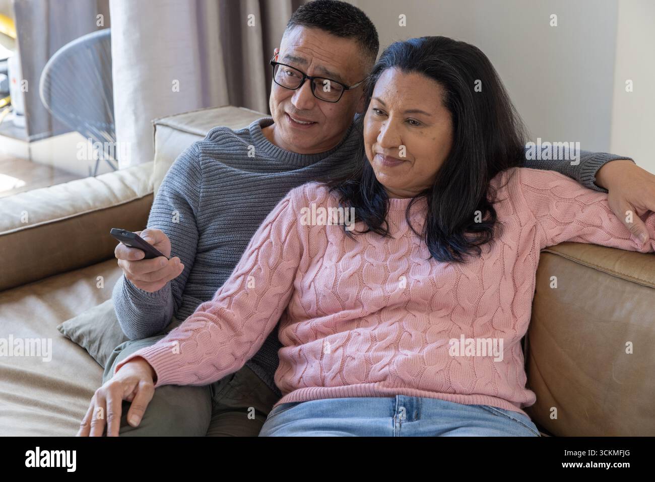 Una coppia senior seduto su un divano in pelle nel soggiorno con telecomando della tv e cuscino di lancio Foto Stock