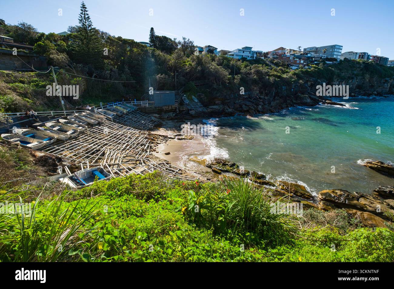 Le barche a remi riposano su scaffali strutturati lungo una costa pittoresca con limpide acque turchesi, circondati da vegetazione verde e strutture urbane, Foto Stock
