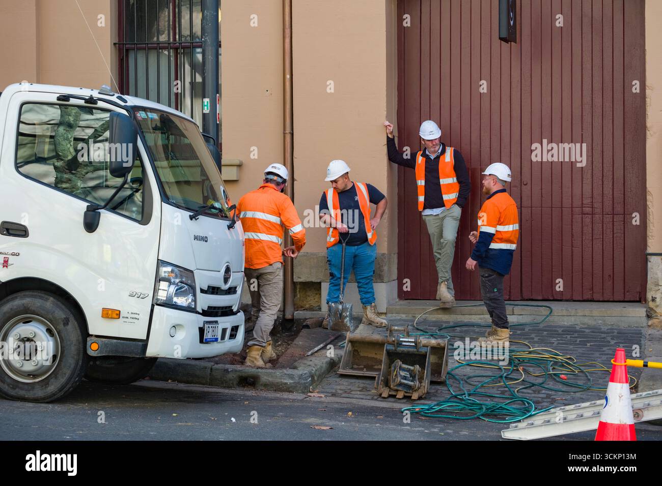 Gli operai edili con giubbotti e caschi ad alta visibilità si riuniscono vicino a un camion e alle attrezzature, impegnandosi in una conversazione. La scenografia include strumenti, Safet Foto Stock