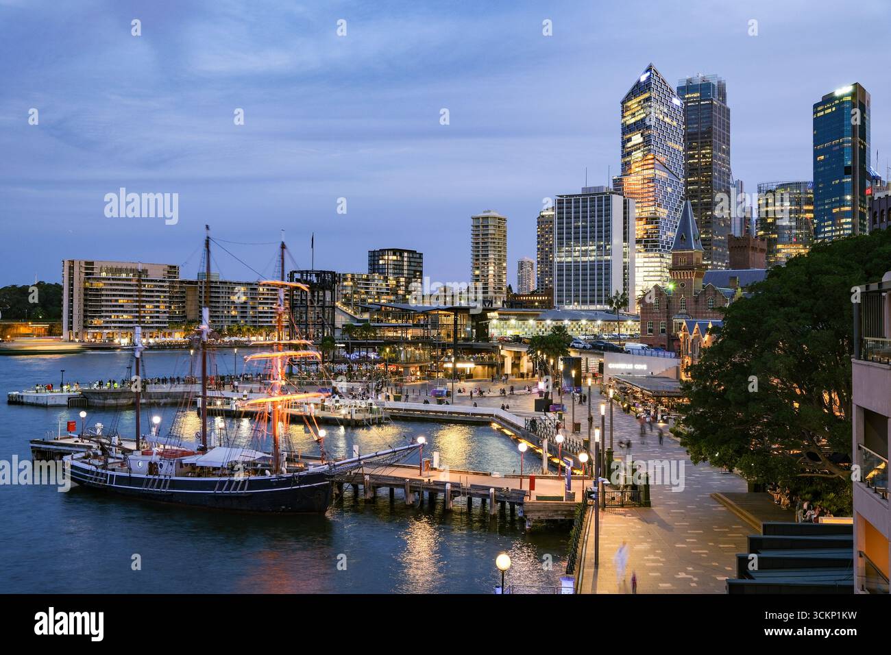Splendido paesaggio urbano serale caratterizzato da un'affollata piazza fronte mare, edifici illuminati, una nave ormeggiata e un vivace ambiente urbano, The Rocks, Sydney Foto Stock