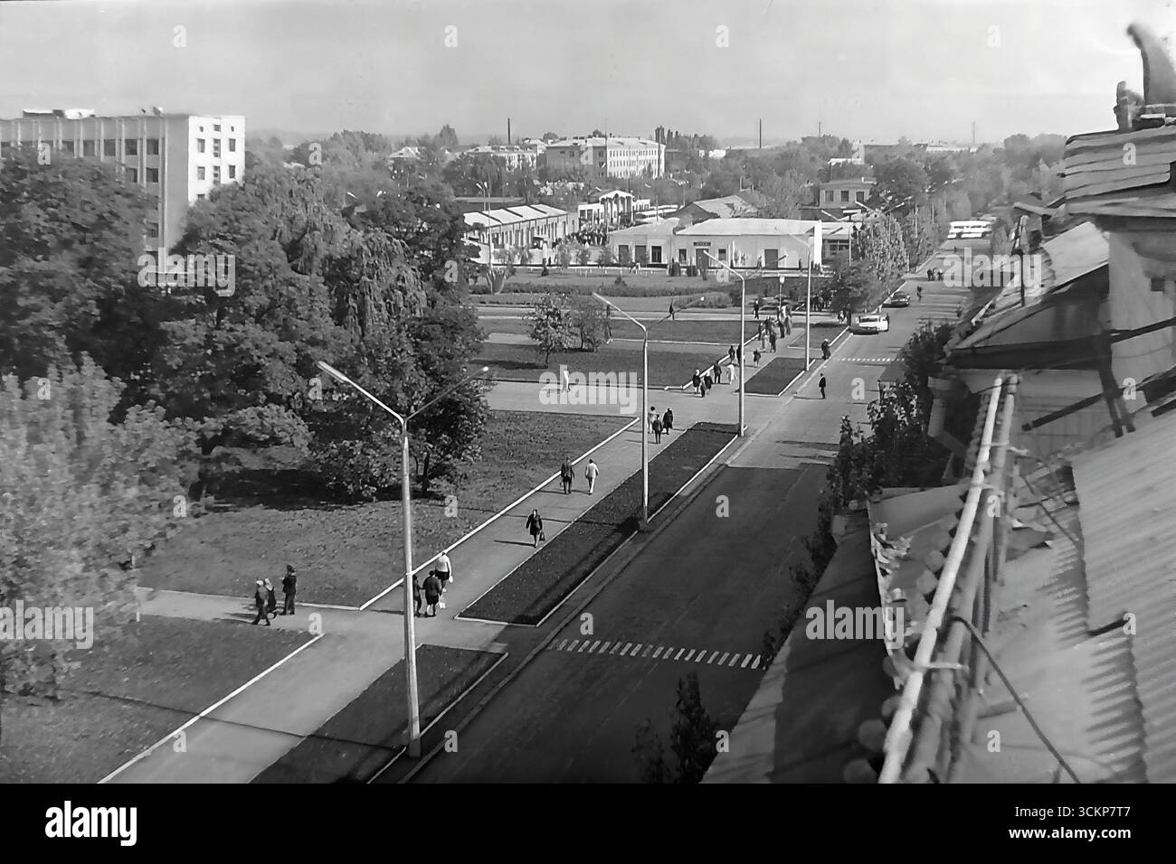 Un viale spazioso vicino alla piazza centrale della Rivoluzione d'ottobre a Sloviansk, 1976. I cittadini passeggiano lungo l'ampio vicolo verde sullo sfondo degli edifici dell'epoca sovietica, tra cui un padiglione pubblico. Questo scatto è un potente simbolo della calma e della ben ordinata vita urbana che esisteva nel Donbas durante l'era della stagnazione, riflettendo lo stile di vita pacifico e misurato di quel tempo, molto prima dei tragici eventi che avrebbero cambiato per sempre la città Foto Stock
