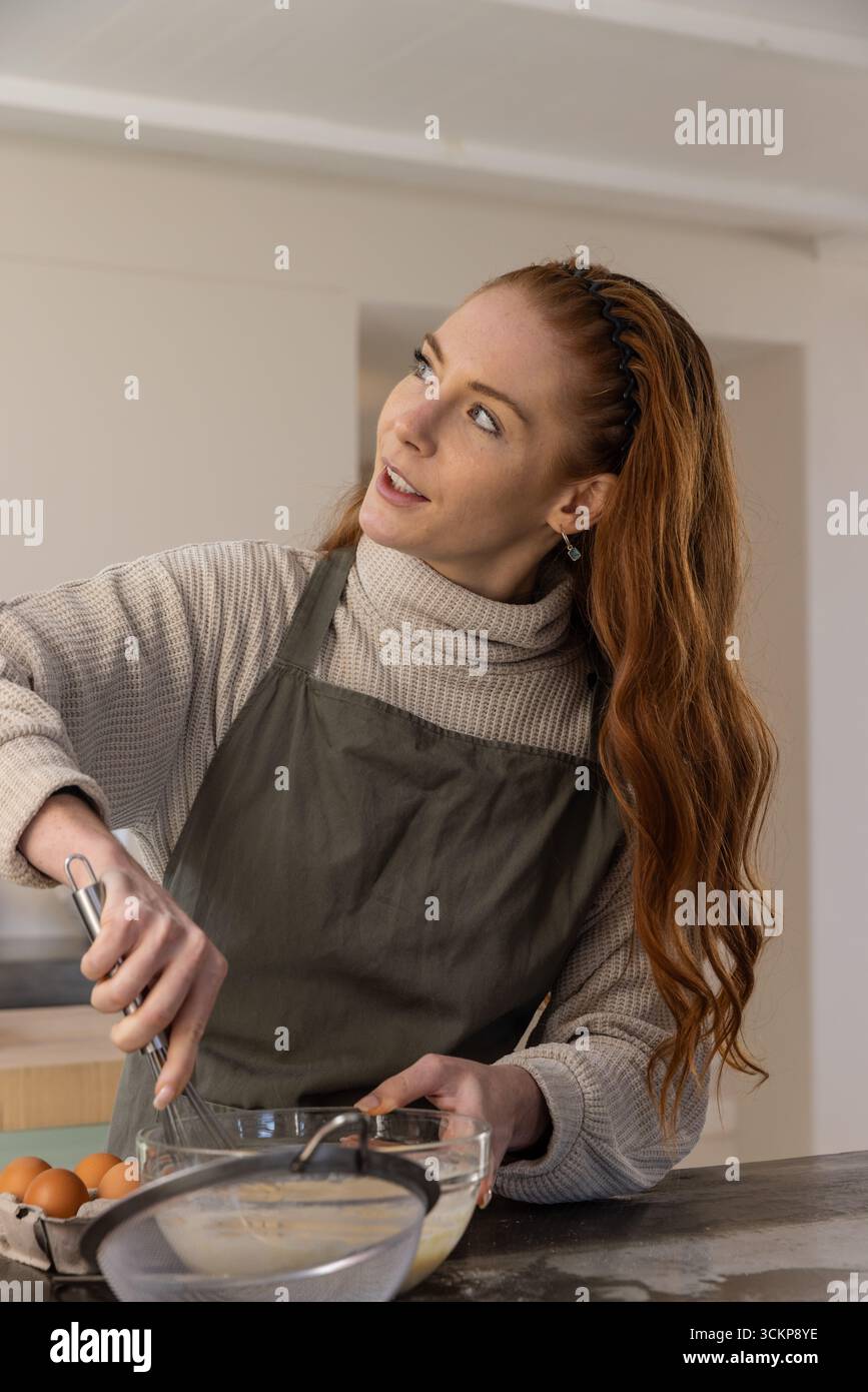 Donna dai capelli rossi che indossa un grembiule con pastella in una ciotola di vetro al banco della cucina Foto Stock