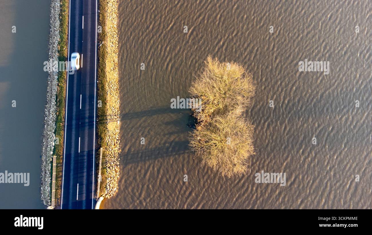Immagine di un drone che mostra un'auto che viaggia su una strada sopraelevata al di sopra dell'acqua alluvionale, con alberi parzialmente sommersi nelle vicinanze Foto Stock