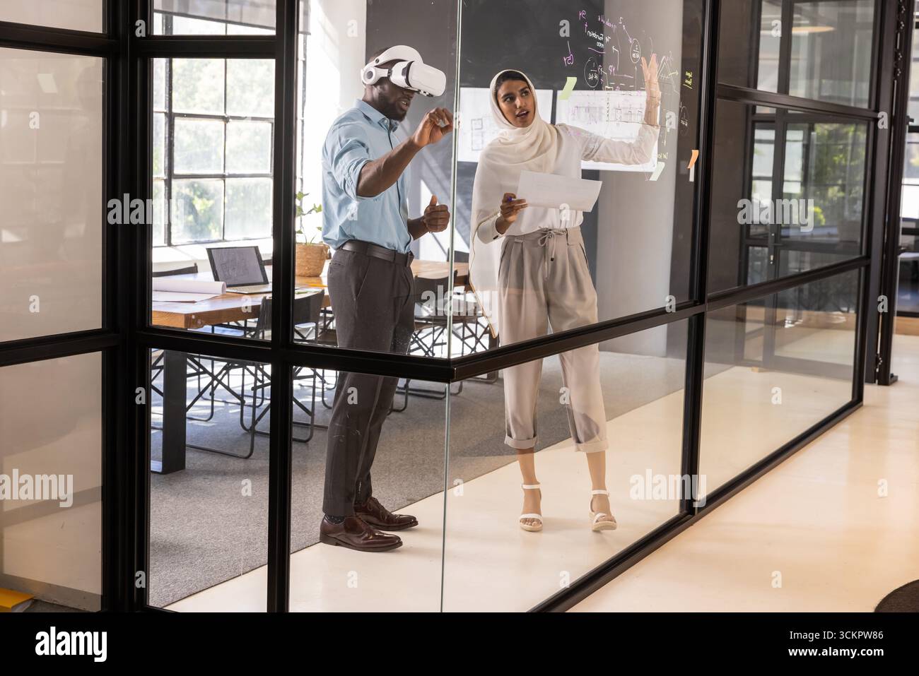 Colleghi diversi in piedi vicino a un pannello di vetro in ufficio, utilizzando attrezzi vr in possesso di piani stampati Foto Stock