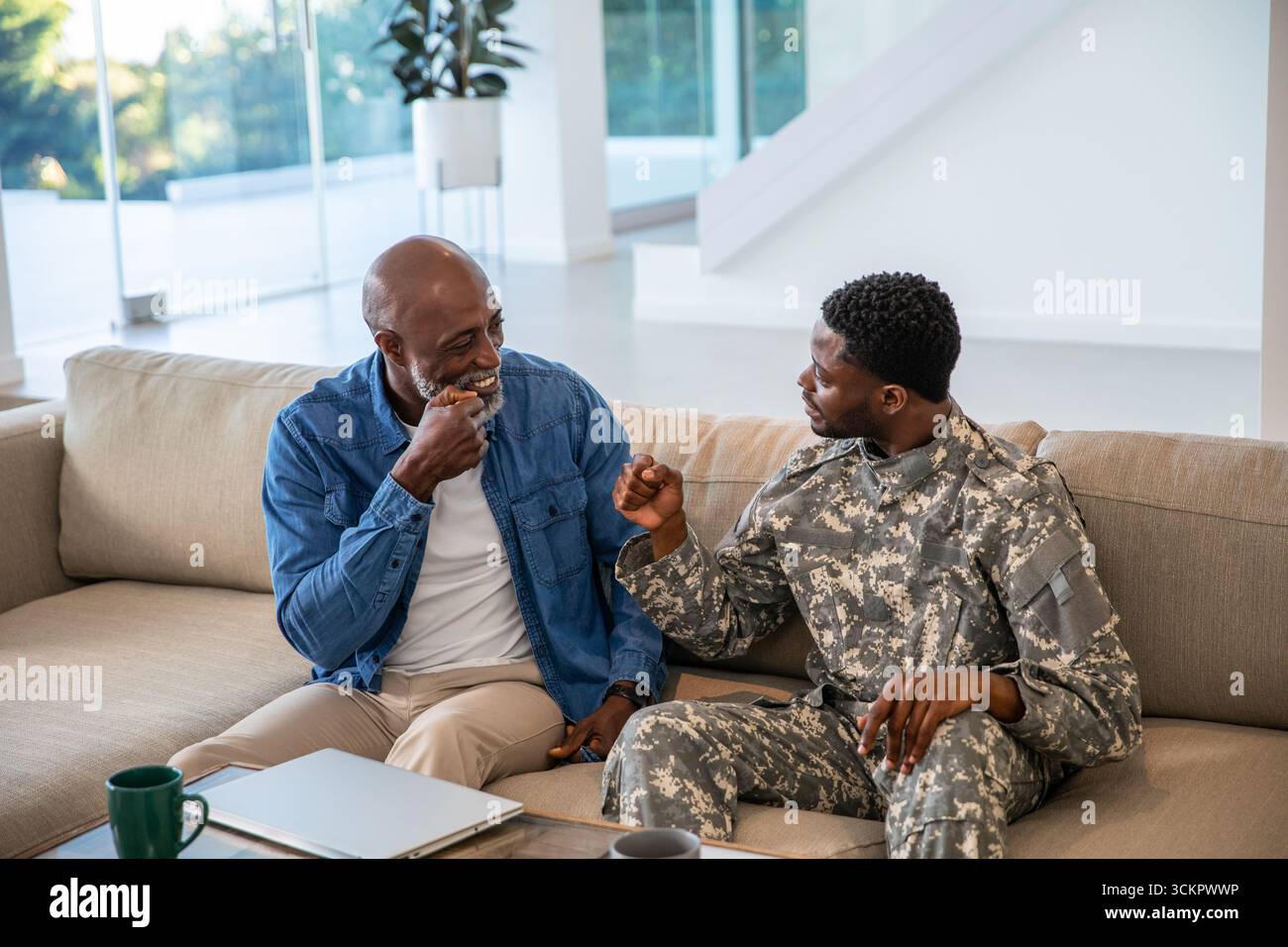 Padre afroamericano che batte pugni con figlio soldato a casa accanto a un tavolino con un computer portatile Foto Stock