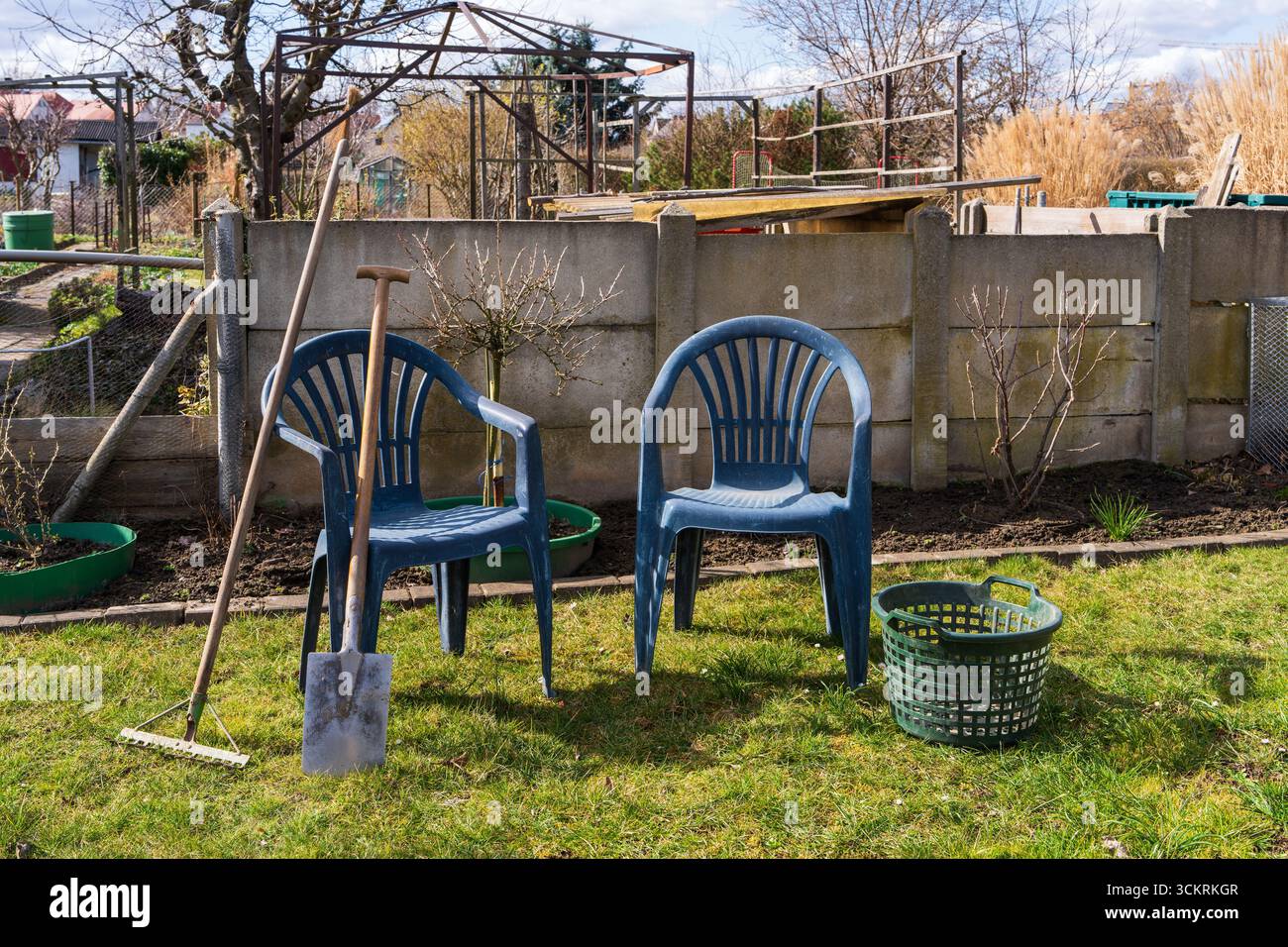 Utensili da giardino accanto a una sedia di plastica su un prato verde in un cortile. Giorno di sole, concetto di giardinaggio, lavoro all'aperto, stile di vita in campagna. Foto Stock