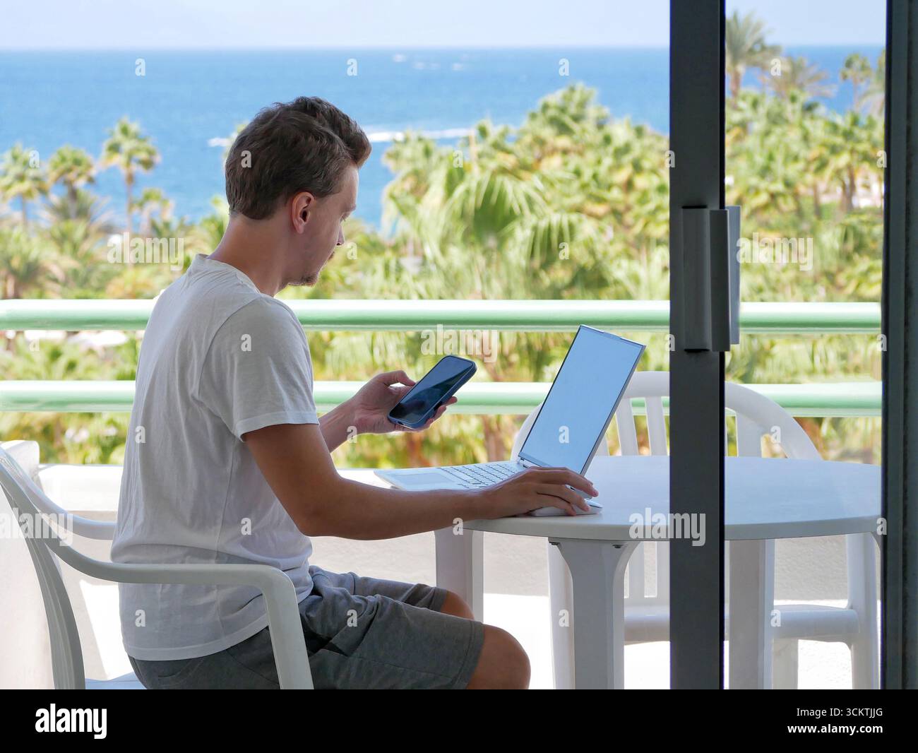 Giovane che lavora a distanza con un computer portatile e uno smartphone su un balcone. Concetto di stile di vita nomade digitale, lavoro freelance, produttività mobile e lavoro Foto Stock