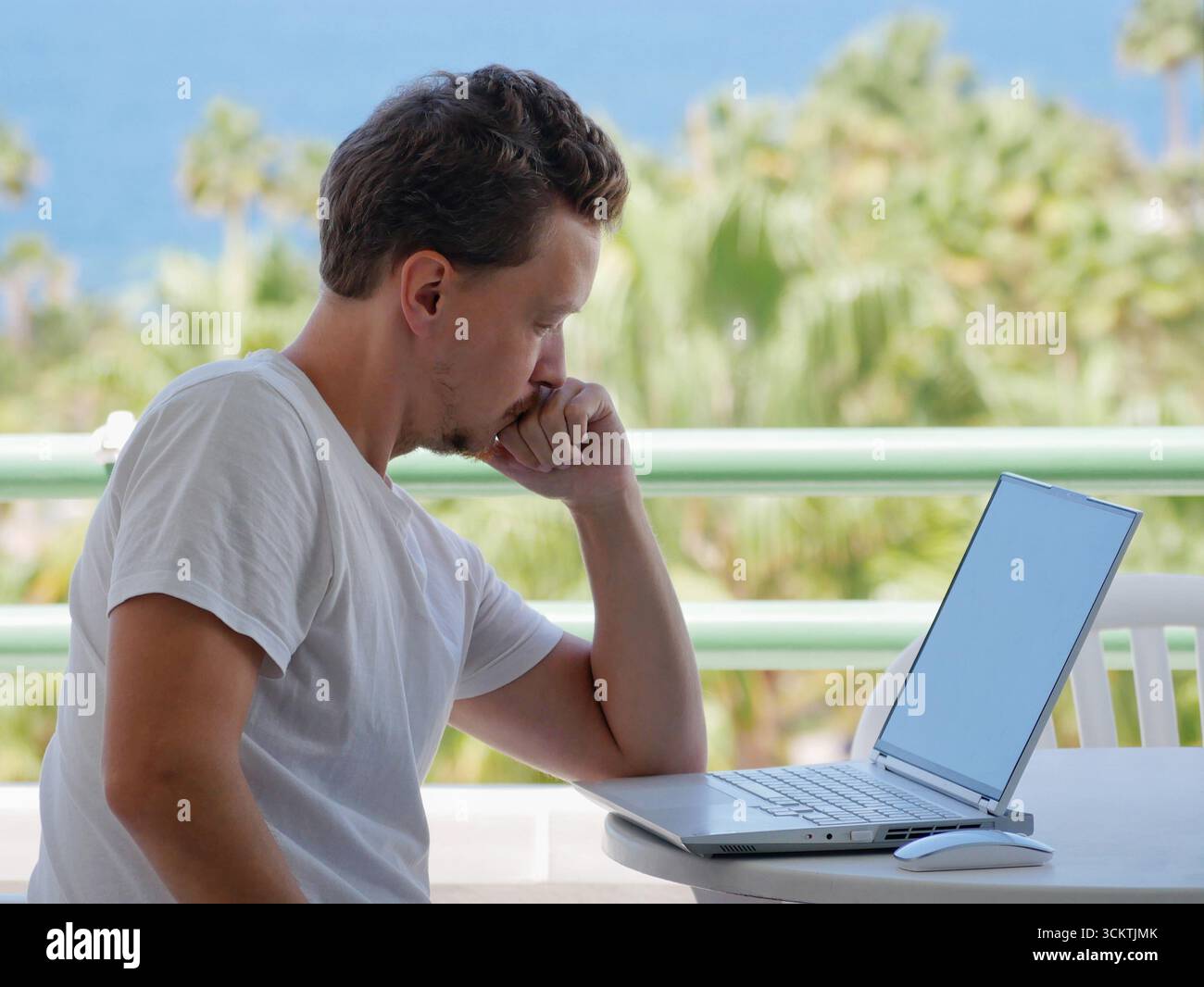 Un giovane attento e premuroso che lavora su un notebook all'aperto con un bellissimo oceano e uno sfondo di palme. Temi di libertà, flessibilità e m Foto Stock