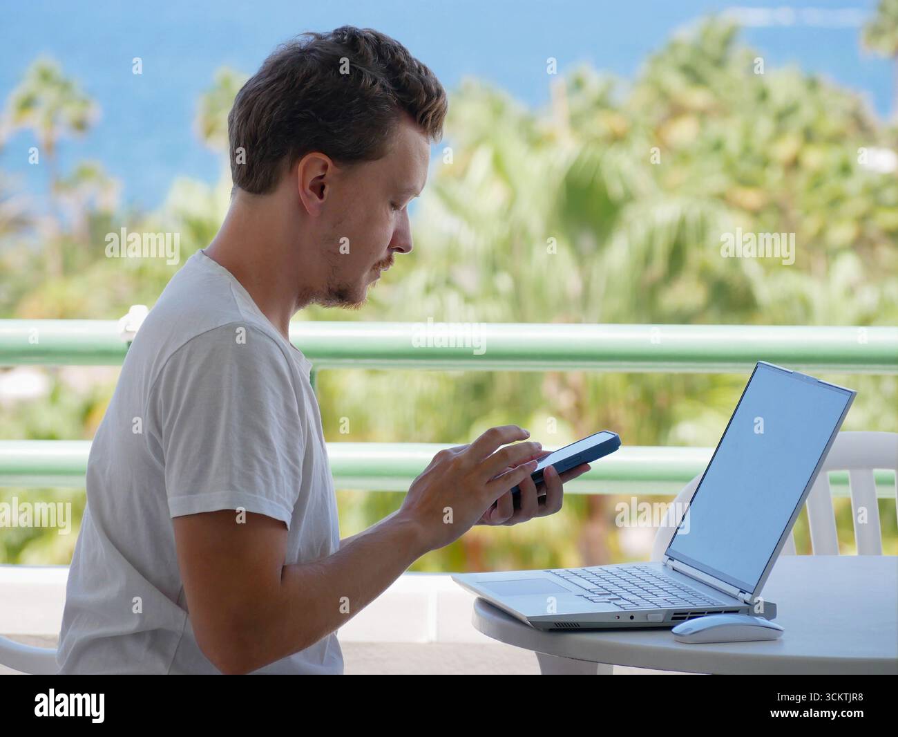 Giovane che lavora a distanza con un computer portatile e uno smartphone su un balcone. Concetto di stile di vita nomade digitale, lavoro freelance, produttività mobile e lavoro Foto Stock