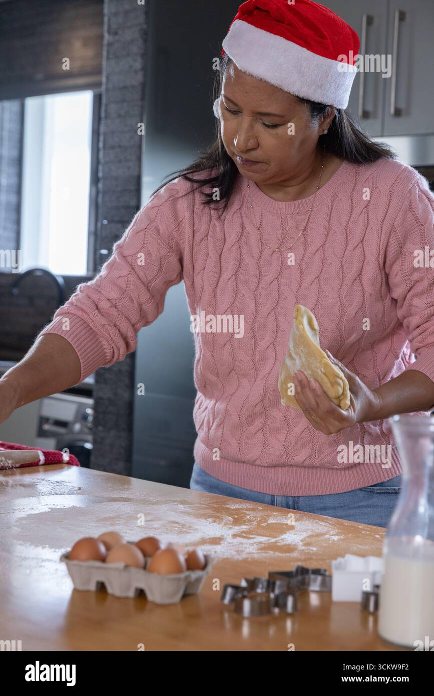 Donna con il cappello di babbo natale che forma l'impasto sul bancone della cucina con farina, taglierini per biscotti, scatola di uova Foto Stock