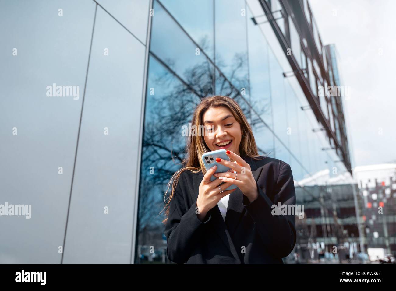 La giovane donna sorride mentre usa il suo smartphone all'aperto in un ambiente urbano con un'architettura moderna che riflette la luce del sole e l'energia vibrante Foto Stock