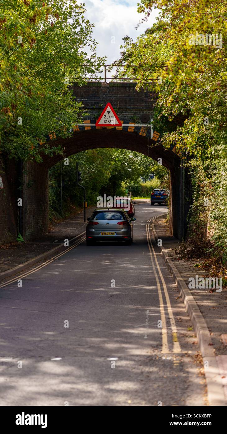 Auto che viaggiano su una strada stretta che si avvicina a un ponte ferroviario basso con cartello di limitazione dell'altezza nel Regno Unito Foto Stock