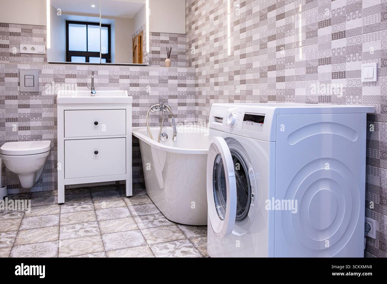 Bagno moderno ed elegante con una funzionale area lavanderia, dotata di lavatrice, vasca elegante e design spazioso con piastrelle chiare e contemporanee Foto Stock