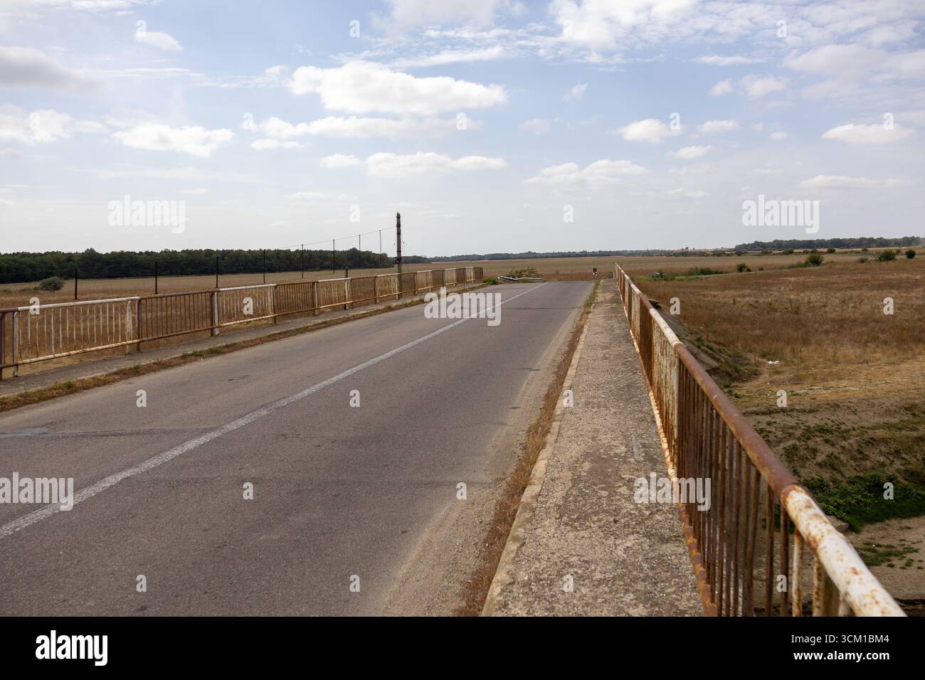 Tranquillo ponte di campagna con una strada asfaltata vuota che si estende attraverso i campi sotto un cielo parzialmente nuvoloso Foto Stock