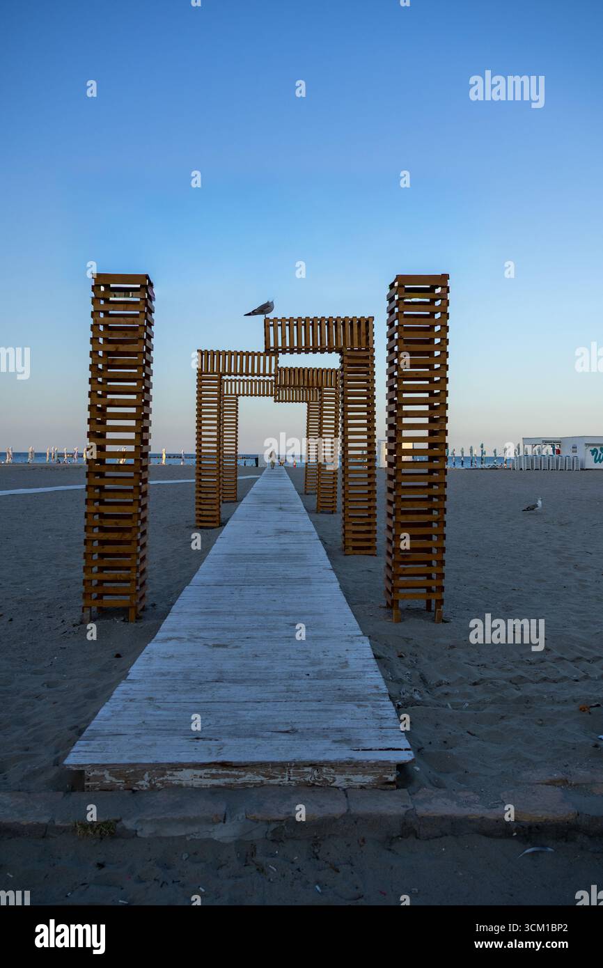 Vista simmetrica di una struttura ad arco in legno sulla spiaggia con un gabbiano arroccato sulla sommità Foto Stock