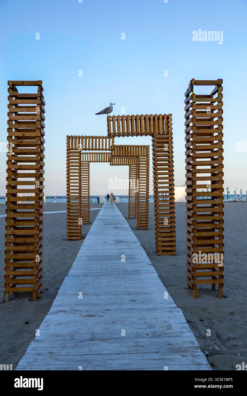 Vista simmetrica di una struttura ad arco in legno sulla spiaggia con un gabbiano arroccato sulla sommità Foto Stock