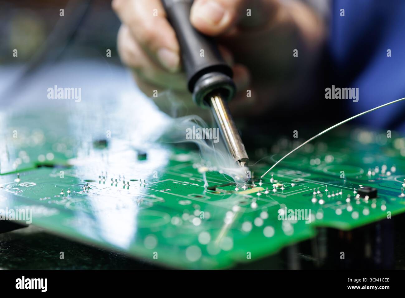 Primo piano della saldatura di un tecnico su un circuito stampato verde con fumo visibile durante l'assemblaggio dei componenti elettronici presso una postazione di lavoro di fabbrica Foto Stock