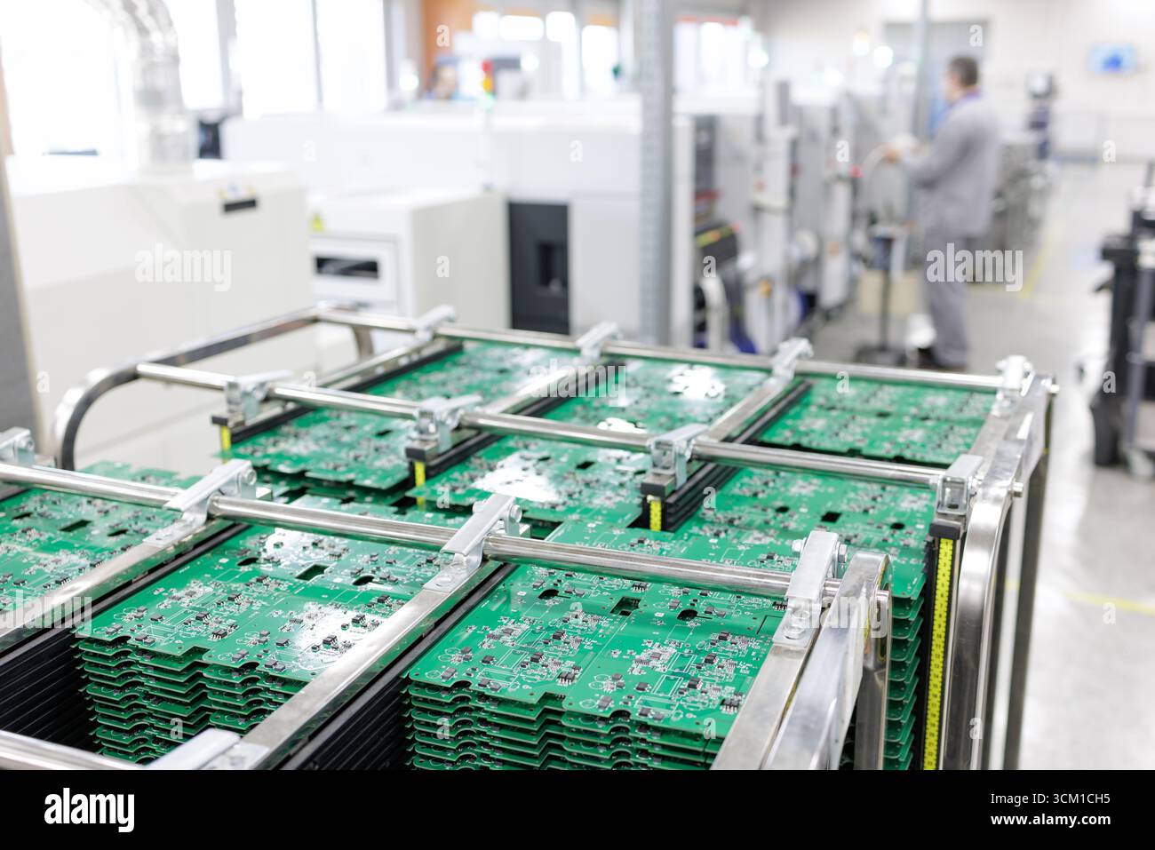 Pannelli per circuito stampato impilati in un rack ESD con tecnici e apparecchiature SMT sullo sfondo su una moderna linea di produzione elettronica automatizzata Foto Stock
