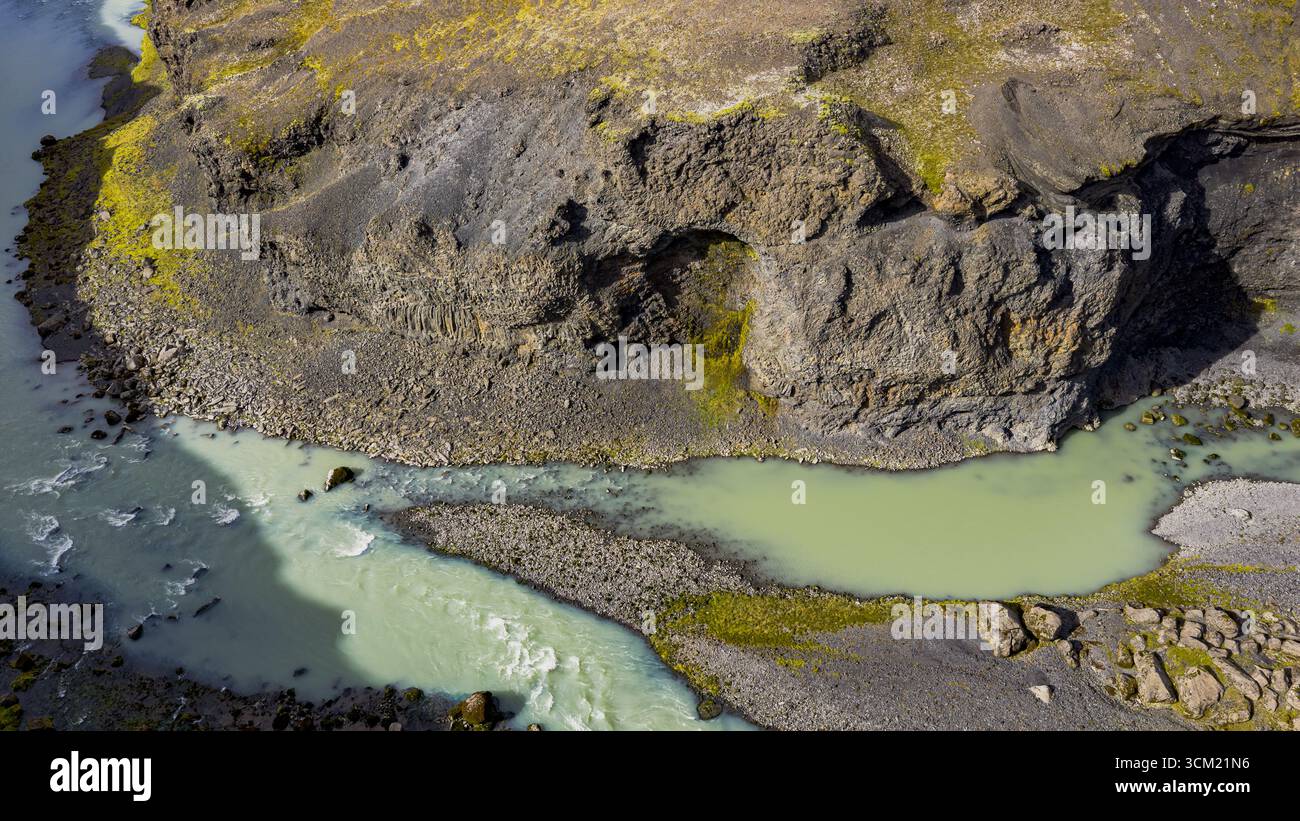 Un tortuoso fiume turchese scorre attraverso formazioni scure di basalto in Islanda. Un arco naturale come la parete rocciosa e il terreno muschiato aggiungono interesse geologico. Foto Stock