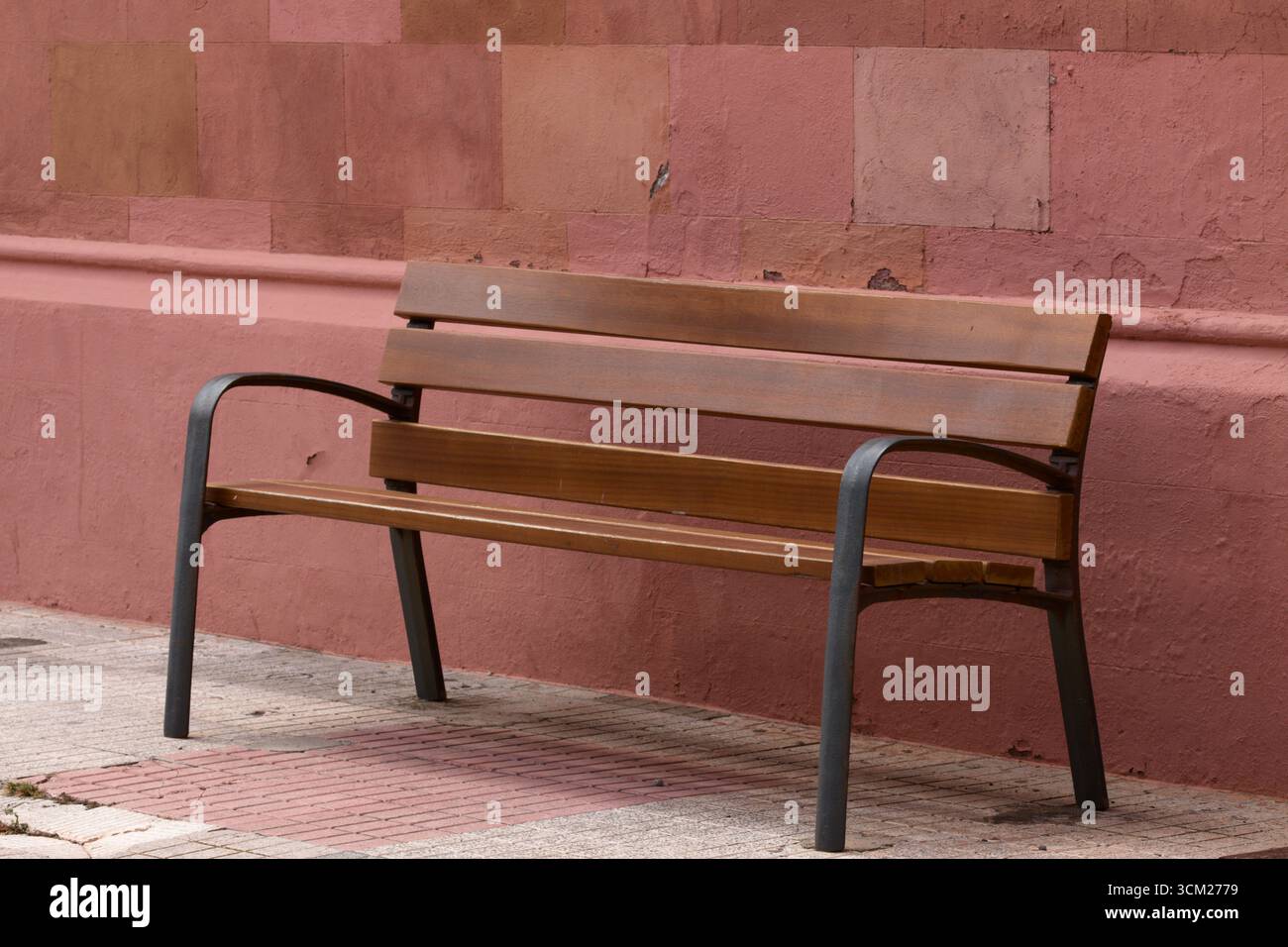 Immagine di una panca di legno in uno spazio pubblico all'aperto accanto a una parete marrone e seduta su un pavimento piastrellato Foto Stock