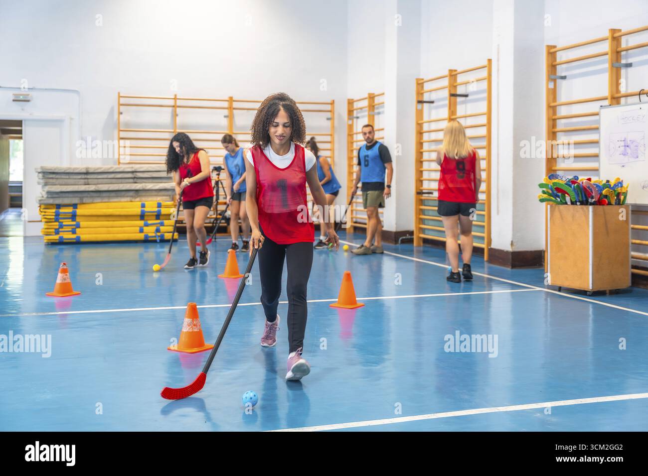 Una giovane donna che pratica hockey da pavimento, dribbling la palla con il bastone, durante una sessione di allenamento in una palestra, circondata da compagni di squadra e allenatori, concentrati Foto Stock
