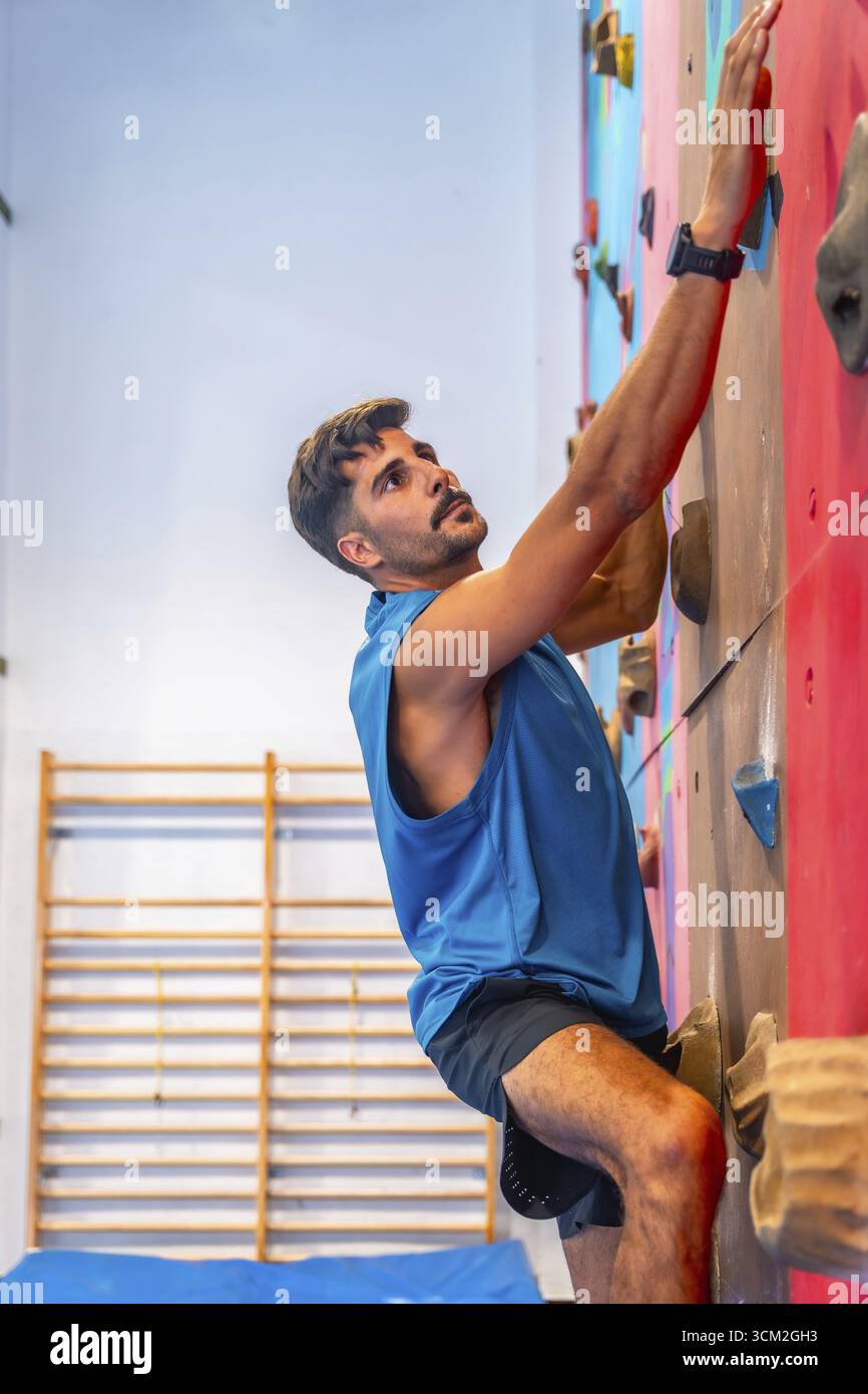 Atleta maschile concentrato, afferrando meticolosamente le maniglie colorate durante l'ascesa di una parete artificiale per arrampicata, dimostrando forza, agilità, Foto Stock