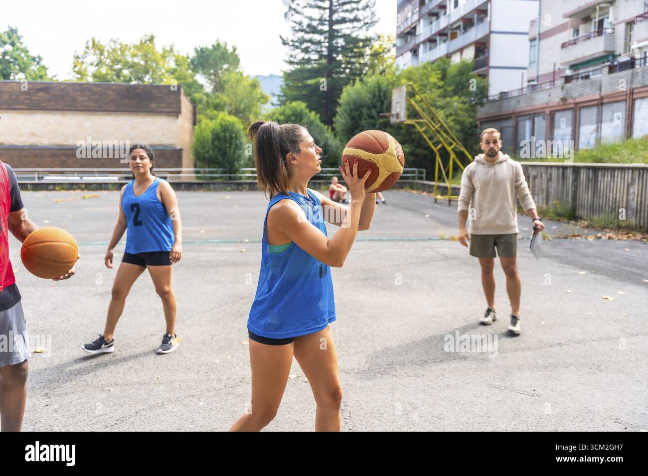Giocatrice di pallacanestro concentrata in uniforme blu durante l'allenamento all'aperto in una giornata di sole, osservata da un allenatore con appunti e squadra Foto Stock