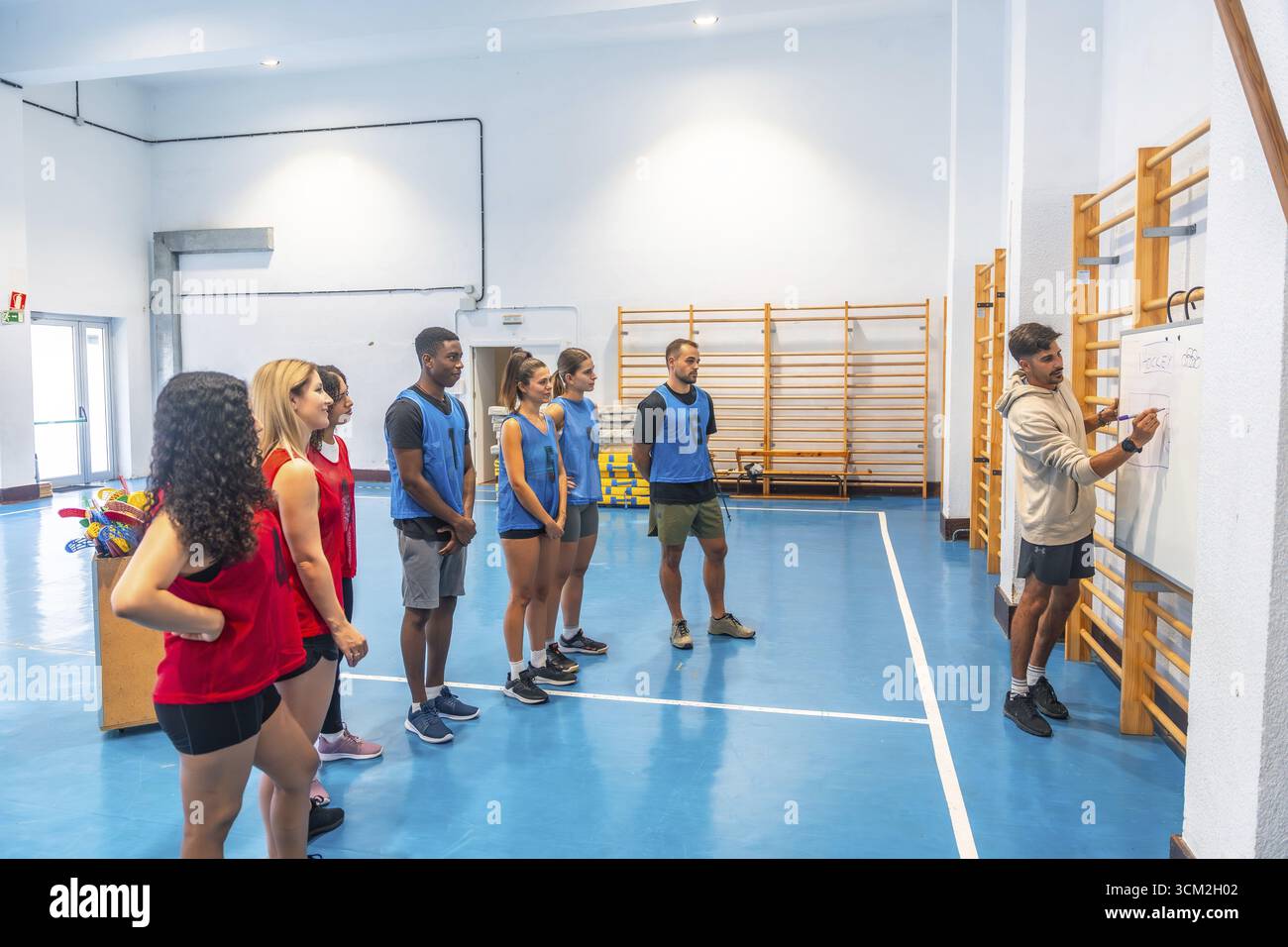 Allenatore di hockey disegnando un piano di gioco sulla lavagna mentre spiega la strategia ai giocatori divisi in due squadre, indossando uniformi rosse e blu, ascoltando Foto Stock