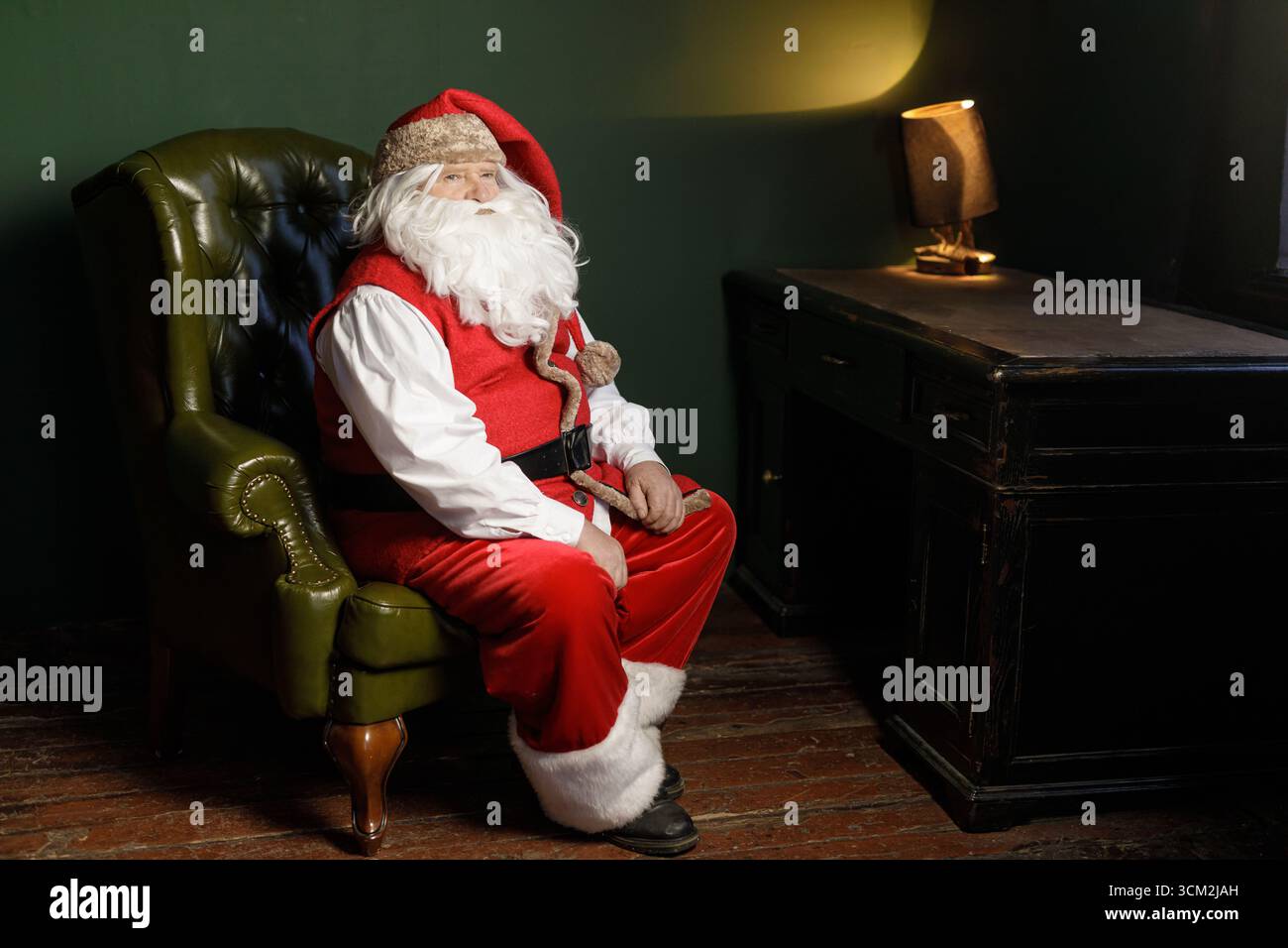 Babbo Natale in tuta rossa si trova accanto a una scrivania e una lampada vintage con ampio spazio per le copie, ideale per annunci natalizi, saluti e campagne stagionali Foto Stock
