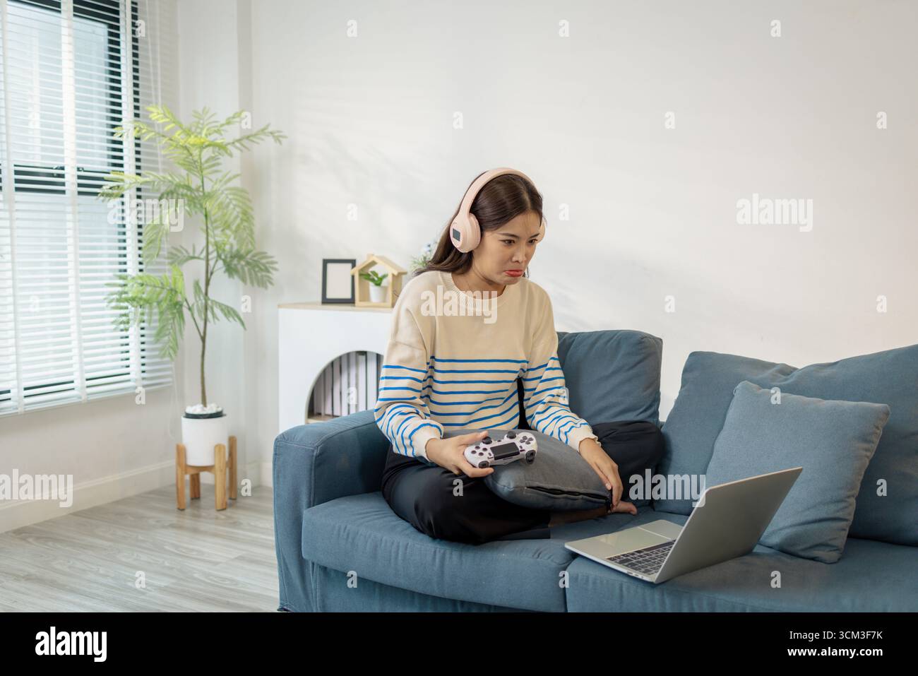 Una giovane donna che indossa le cuffie sta giocando a un videogame sul suo notebook, tenendo in mano un controller con espressioni emotive di gioia, eccitazione e fruste Foto Stock