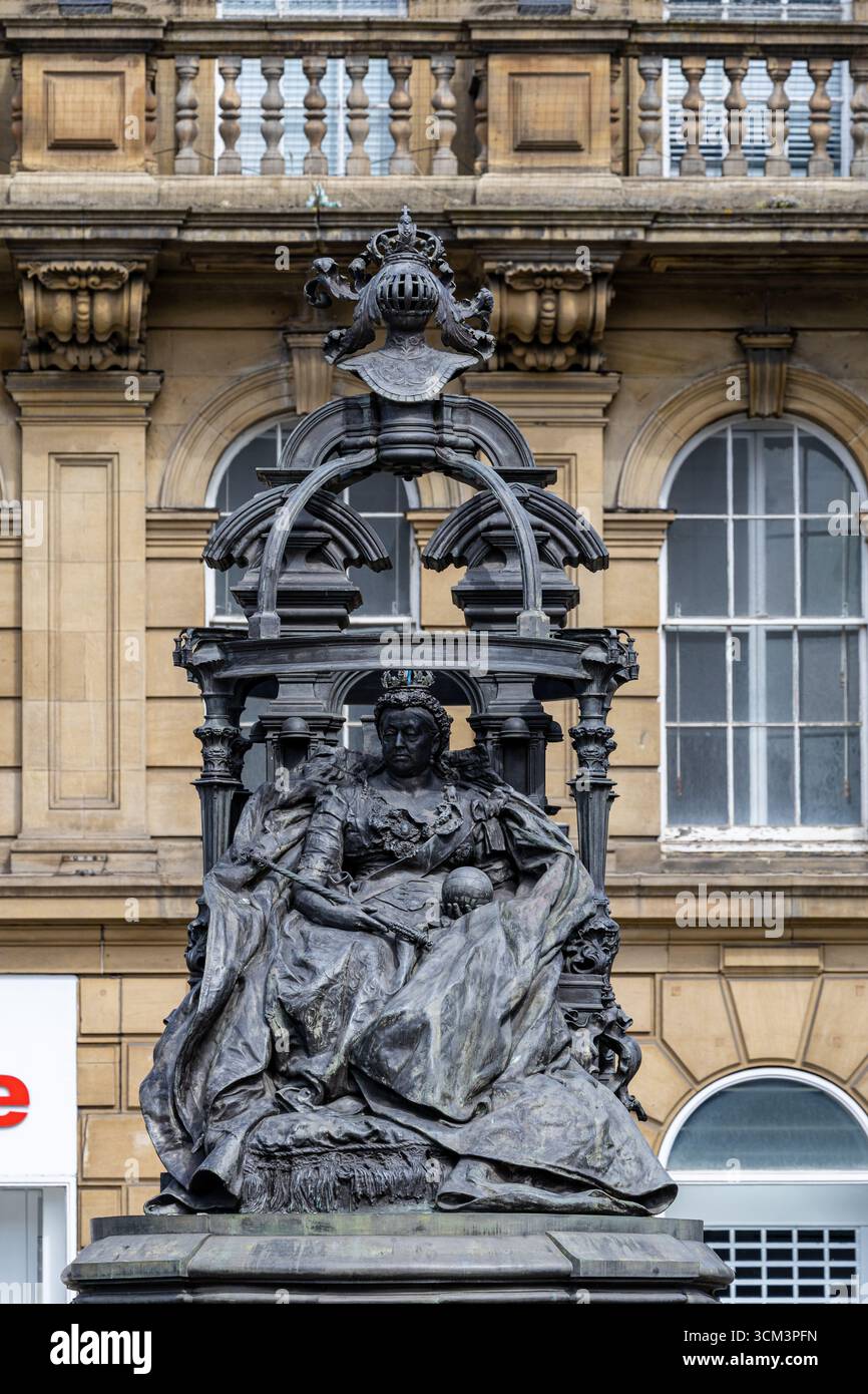 Una dettagliata statua in bronzo della regina Vittoria, seduta su un trono, è incorniciata centralmente sullo sfondo di un classico edificio in pietra con Windo ad arco Foto Stock