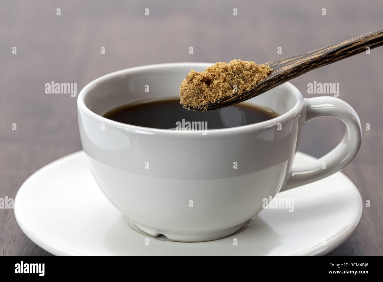 Un caffè nero con cucchiaio pieno di zucchero di canna, tazza e piattino in ceramica bianca su un tavolo di legno, bevanda mattutina, atmosfera calda e invitante Foto Stock