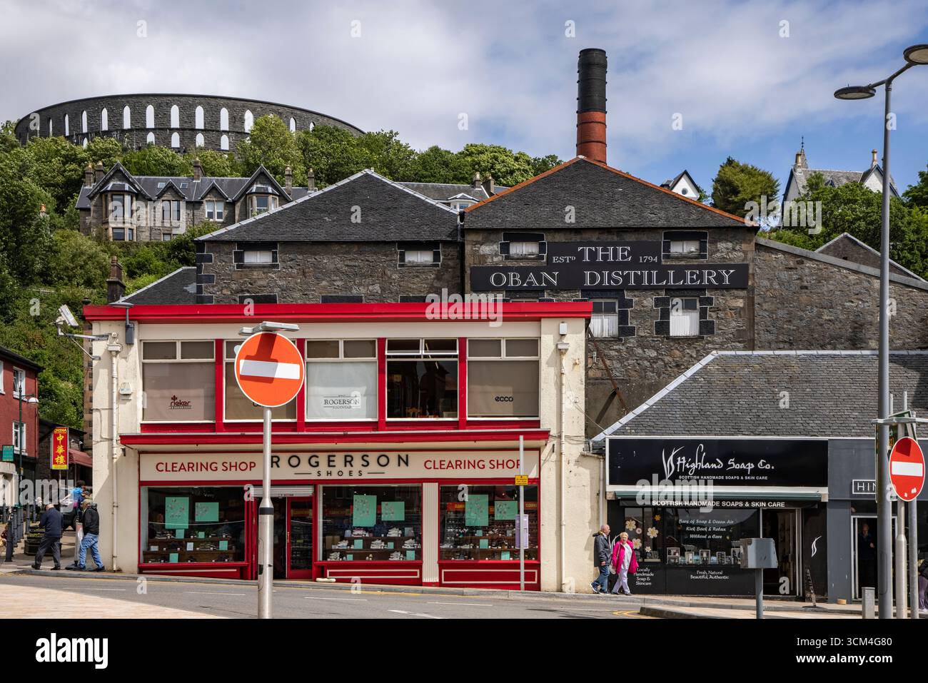 Negozi nel centro della città con la distilleria Oban e la McCaig'S Tower (o McCaig'S Folly) Behind, Oban, Scotland, Argyll and Bute Council, United K Foto Stock