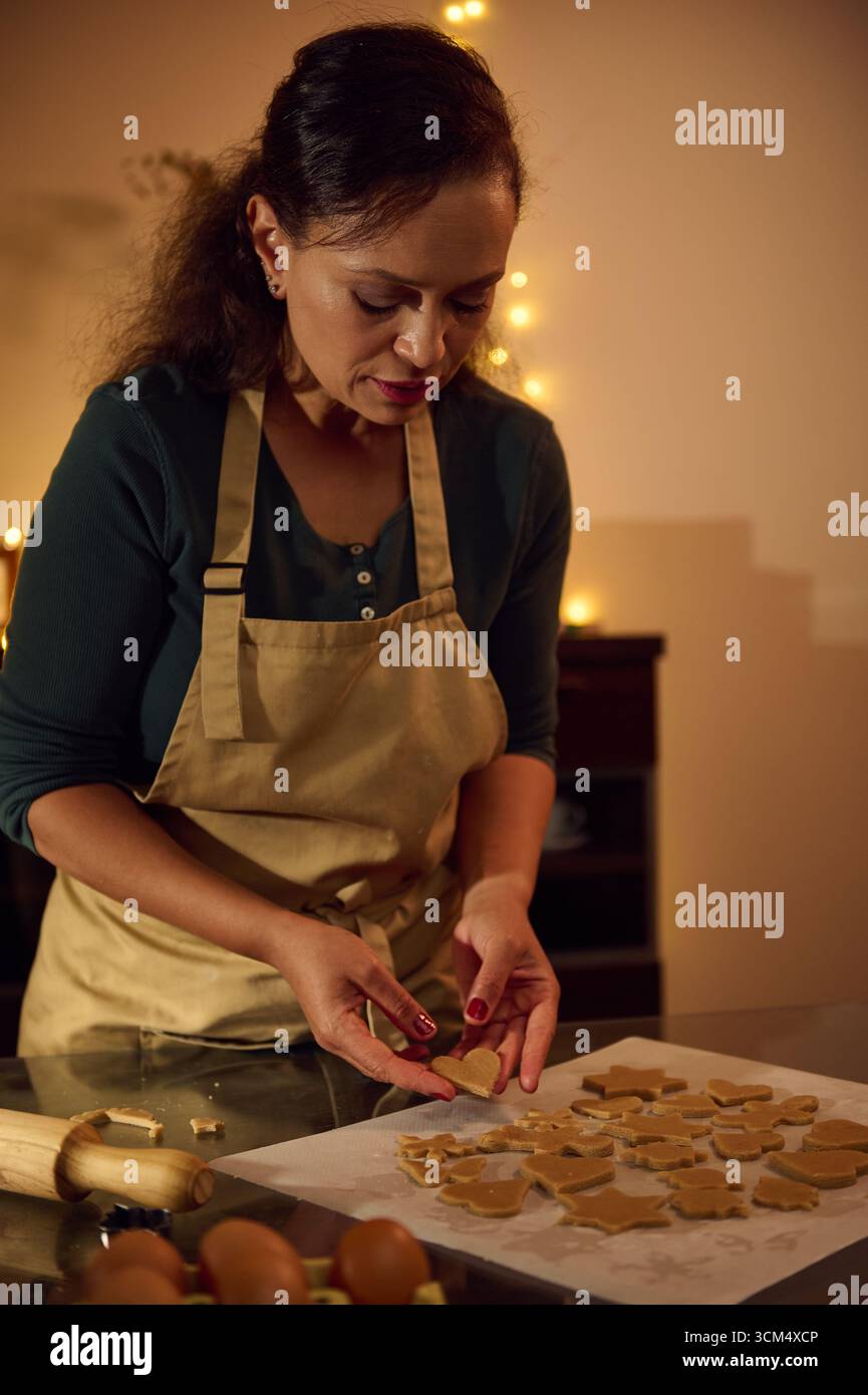 Una donna che indossa un grembiule forma biscotti di pan di zenzero in una cucina calda illuminata decorata per le vacanze. La scena evoca un'atmosfera accogliente e festosa Foto Stock