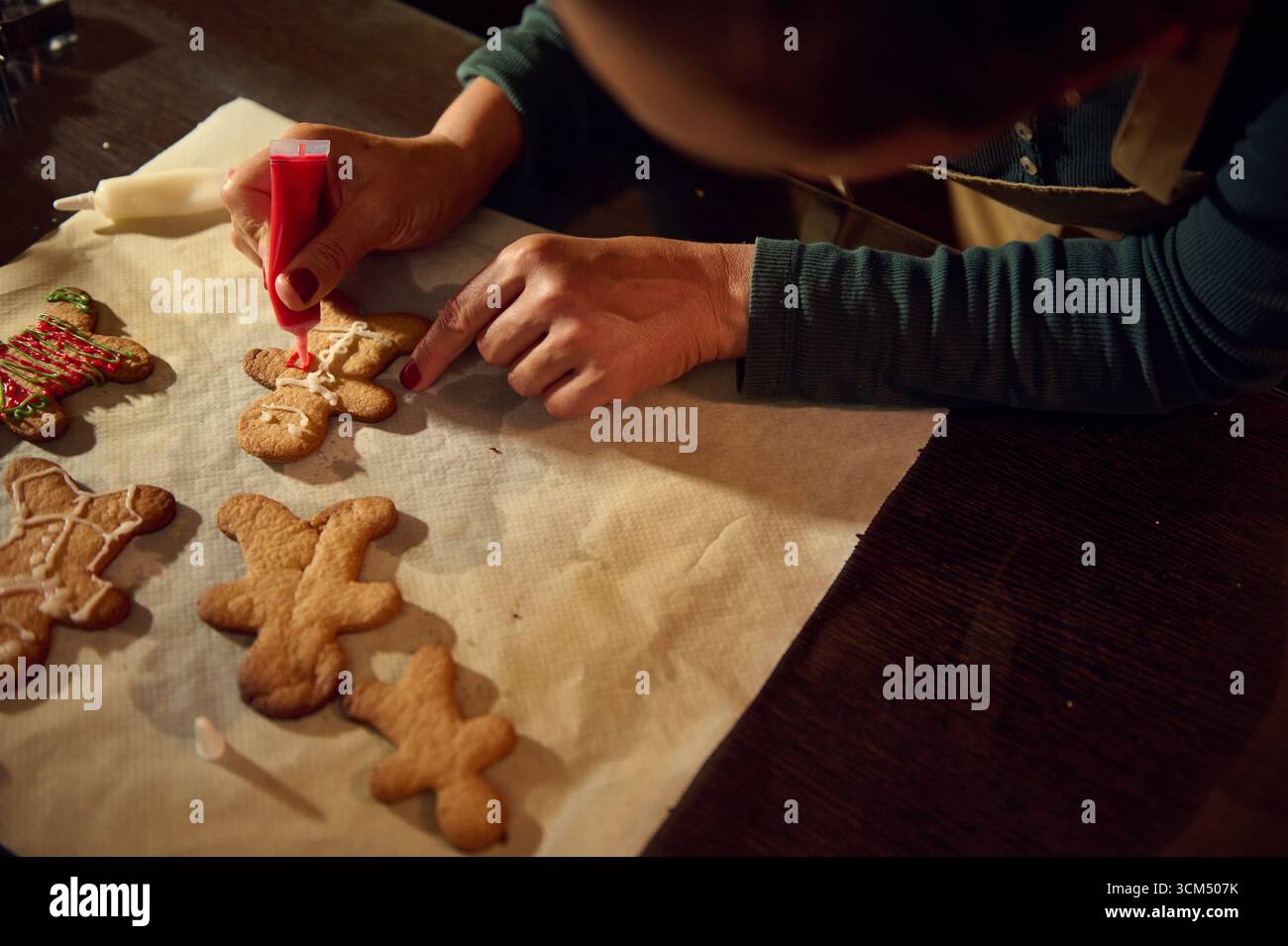 Una donna con un tubo di pasticceria decora i biscotti al pan di zenzero a forma di gente durante i preparativi natalizi. Il calore e la creatività delle vacanze riempiono lo scen Foto Stock