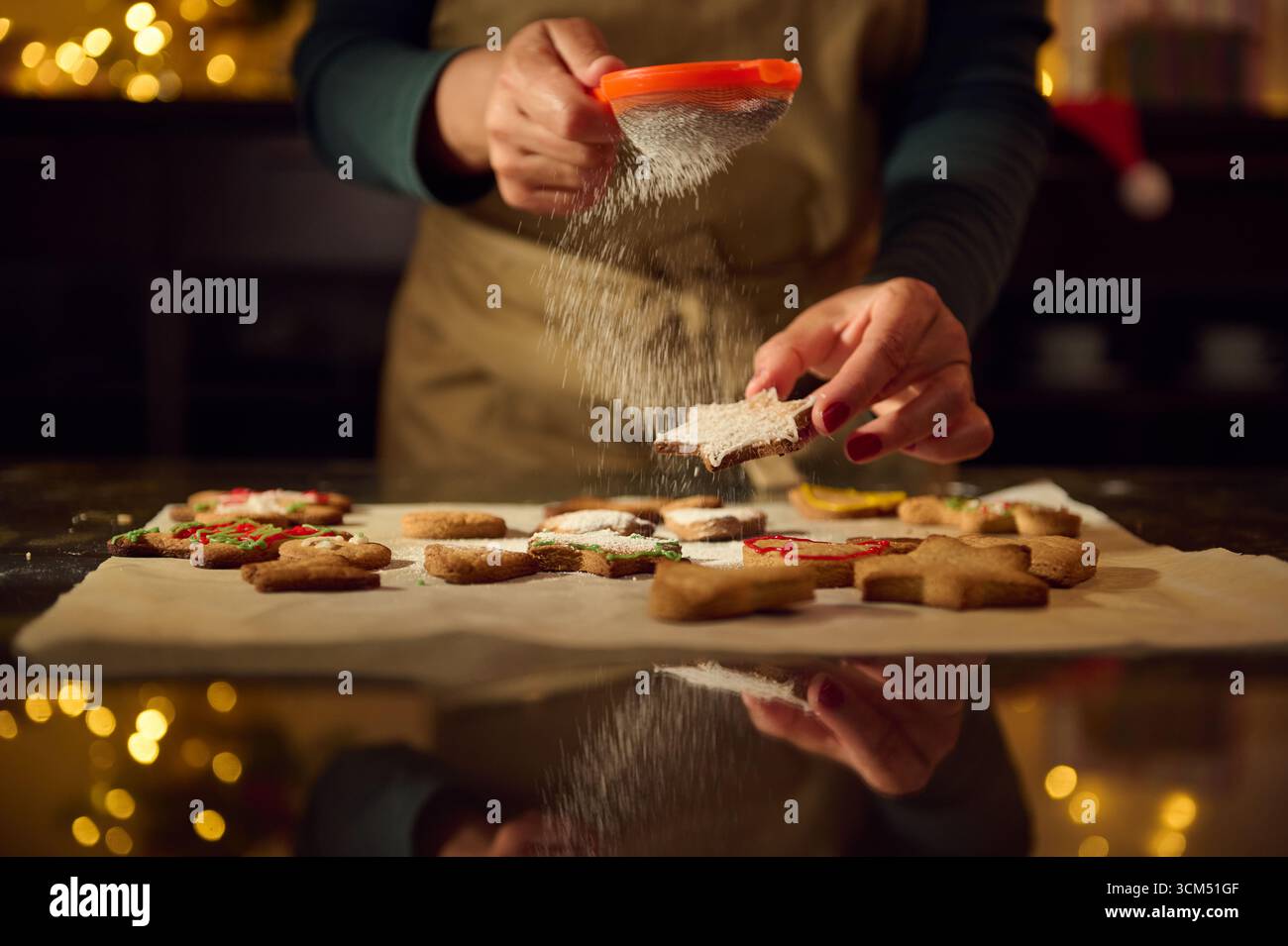 Una persona cosparge di zucchero a velo su biscotti appena sfornati a forma di stella. La cucina è illuminata da luci natalizie, sottolineando un ambiente accogliente e accogliente Foto Stock