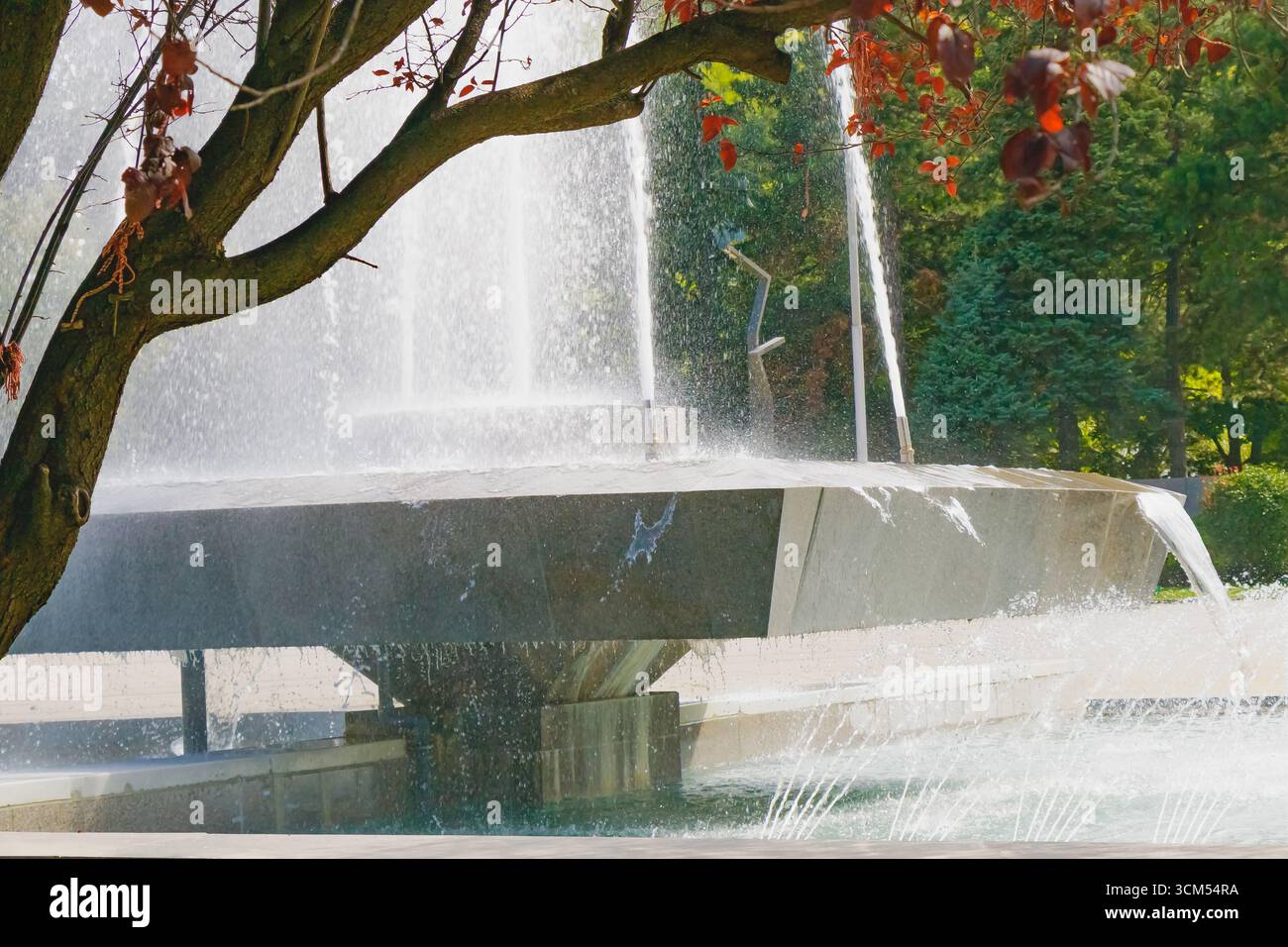Una moderna fontana cittadina con forti getti d'acqua e ruscelli a cascata, incorniciata da rami d'albero con foglie rosse. La scena combina design urbano e natu Foto Stock