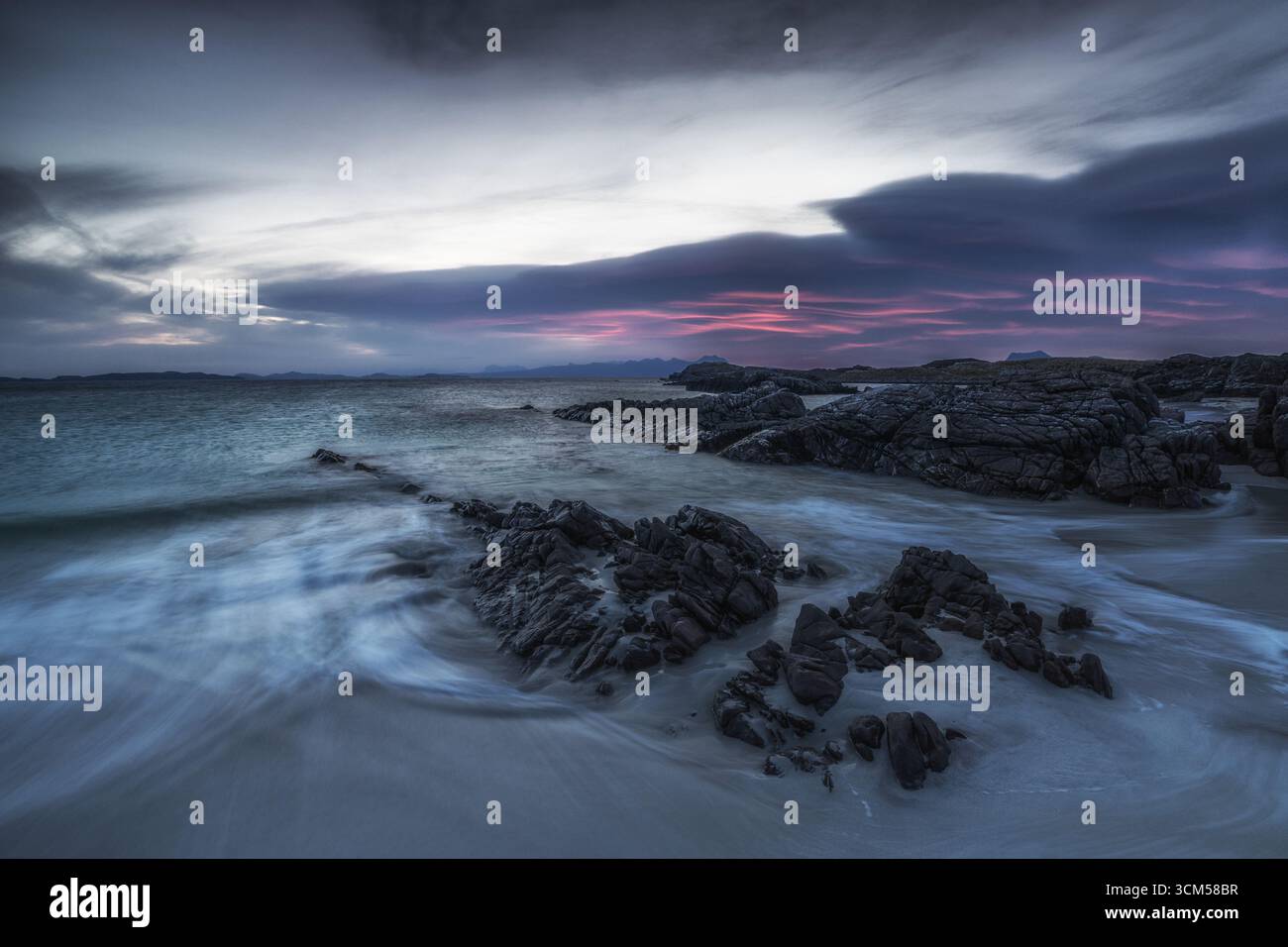 Da una piccola spiaggia di sabbia si tuffano nel mare delle rocce. Le nuvole nel cielo si illuminano di rosso dal basso. Foto Stock